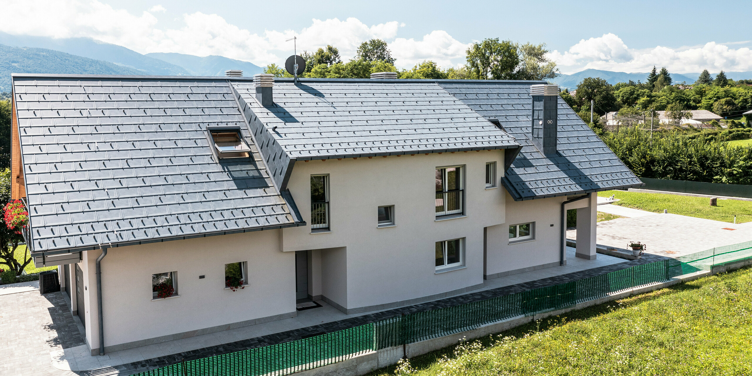 Ein Einfamilienhaus in Belluno, Italien, mit einem eleganten PREFA Dach aus R16 Dachplatten in P.10 anthrazit. Das Dachentwässerungssystem, bestehend aus einer Kastenrinne und einem Drainagerohr, ist farblich abgestimmt und vervollständigt das ästhetische und funktionale Design des Daches. Mit seiner traditionellen Bauweise und dem modernen Dachmaterial bildet das Haus einen reizvollen Kontrast zur malerischen Bergkulisse.