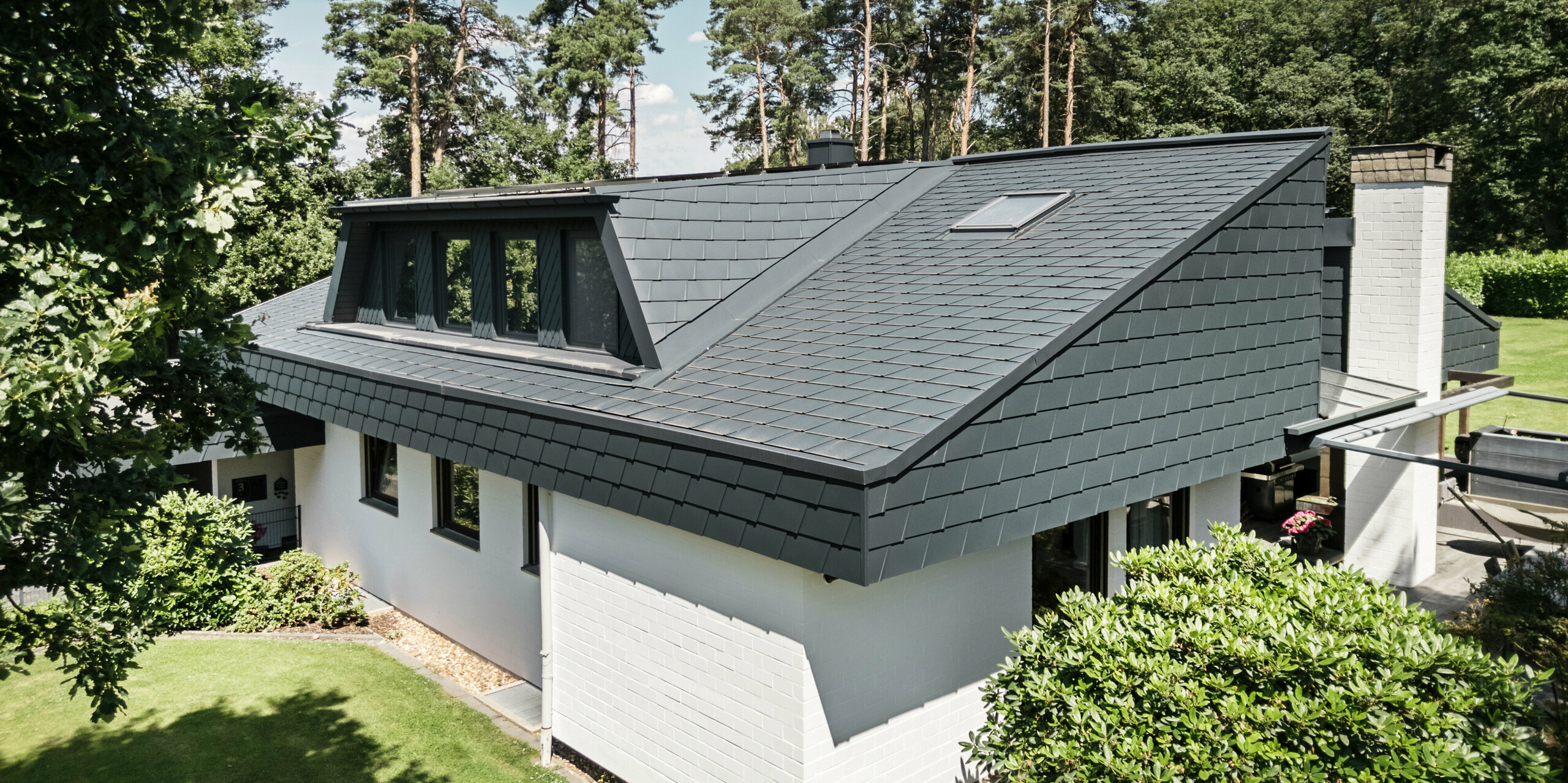 Vogelperspektive auf ein Einfamilienhaus in Mechernich, Deutschland, mit PREFA Dachschindeln DS.19 und einer verkleideten Trapezgaube in P.10 Anthrazit. Das Dach ist mit langlebigen Aluminium-Dachschindeln gedeckt, die eine wetterfeste und ästhetische Lösung bieten. Die weiße Fassade des Hauses harmoniert mit der dunklen Dacheindeckung, während Bäume und Büsche die natürliche Umgebung des Hauses ergänzen.