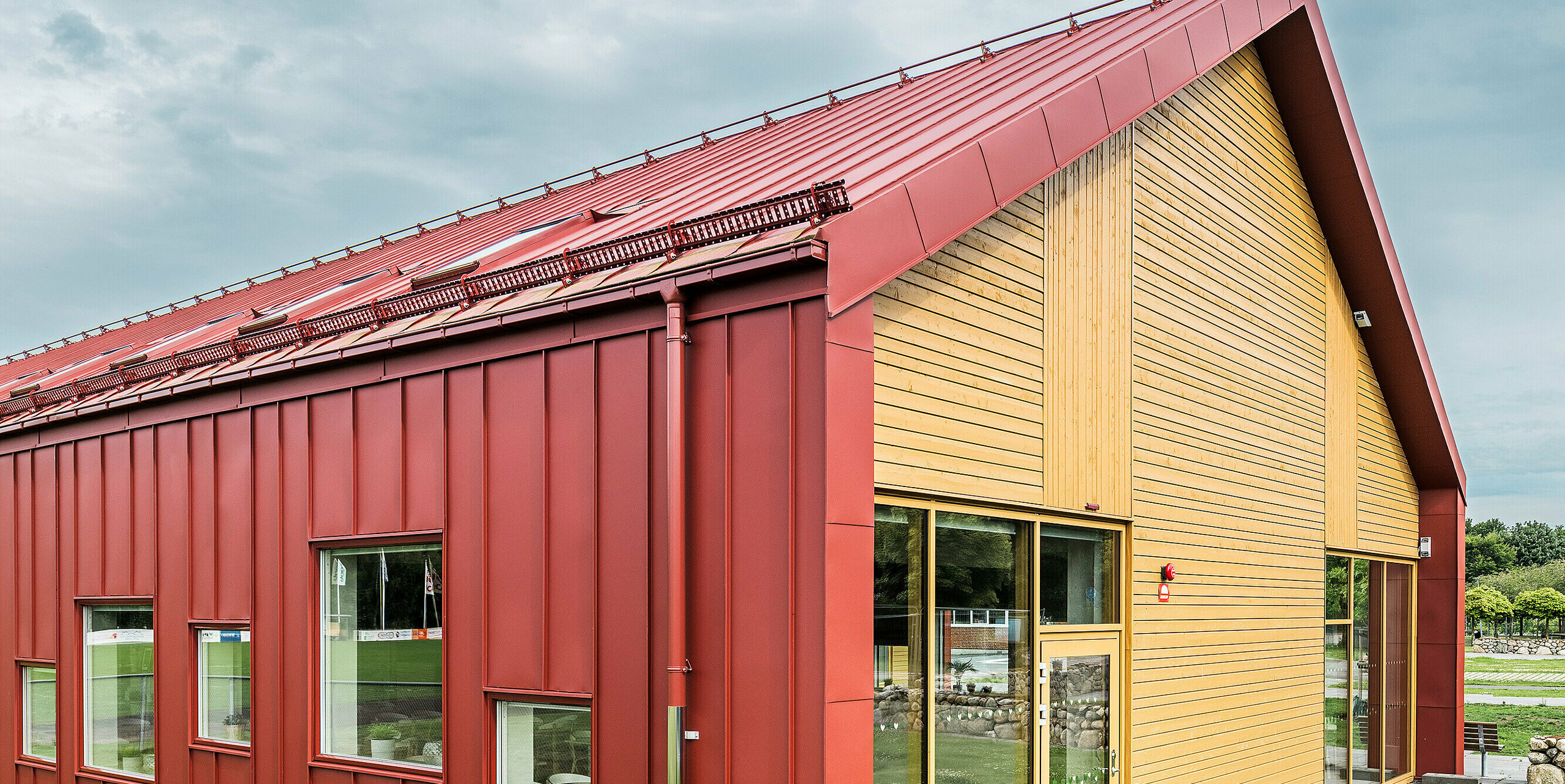 Seitenansicht der Schulkantine Restaurang Gröna Hjärtat in Broby, Schweden. Das Bild zeigt den Giebel des Gebäudes mit einer auffälligen Kombination aus PREFALZ Aluminiumverkleidung in Oxydrot und einer warmen Holzfassade. Die klaren, modernen Linien der Architektur und die Farbkontraste zwischen der roten Stehfalzdeckung und dem natürlichen Holz betonen das innovative Design des Gebäudes. Die Fenster und die Eingangstür auf dieser Seite lassen viel natürliches Licht in die Räume und tragen zur einladenden Atmosphäre bei. Die langlebige Konstruktion aus hochwertigem Blech unterstreicht die Qualität und Nachhaltigkeit des Bauwerks.