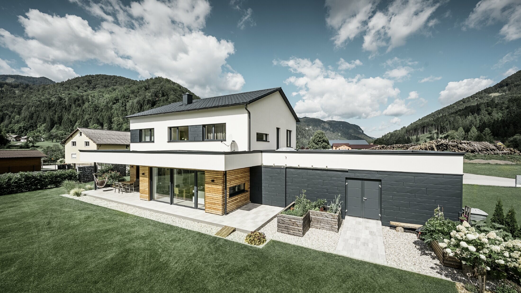 Das Bild zeigt ein modernes Einfamilienhaus in einer idyllischen Landschaft mit einer klaren Sicht auf bewaldete Hügel im Hintergrund. Das Haus zeichnet sich durch eine Kombination aus weißen Putzwänden und einer markanten Fassadenverkleidung mit PREFA Siding.X in P.10 Anthrazit aus, die für einen modernen und langlebigen Look sorgt. Die dunkle Färbung der Fassadenelemente bildet einen ansprechenden Kontrast zum hellen Außendesign und zur natürlichen Umgebung. Die Anordnung der Siding.X Fassadenpaneele ist präzise und verleiht der Immobilie einen strukturierten und hochwertigen Charakter, der typisch für PREFA Produkte ist. Das Dach des Gebäudes wurde mit der bewährten PREFA Dachplatte eingedeckt. Die Dachentwässerung erfolgt durch das Ablaufrohr und die Hängerinne von PREFA.