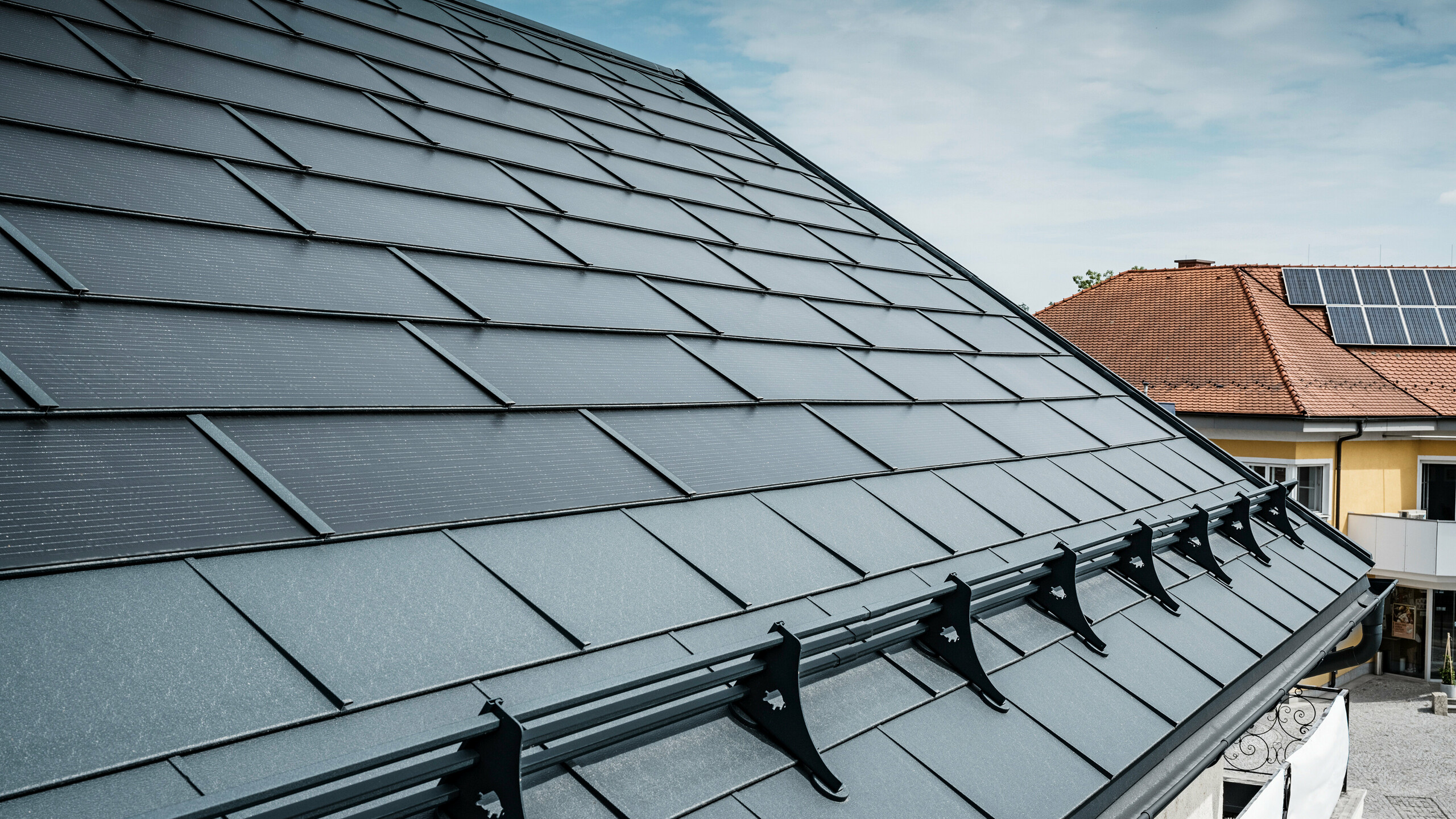 Vy över ett modernt tak med antracitfärgade PREFA solar takplatta. Vid takfoten har taket försetts med klassiska PREFA R.16 takpannor och ett PREFA snöskyddssystem. I bakgrunden syns taket på ett grannhus med traditionella takpannor och solcellsmoduler på taket, vilket understryker kontrasten mellan den nya och den traditionella takkonstruktionen. PREFAs innovativa solcellstakpanel kombinerar estetik och funktionalitet och erbjuder en hållbar energilösning för alla typer av byggnader.