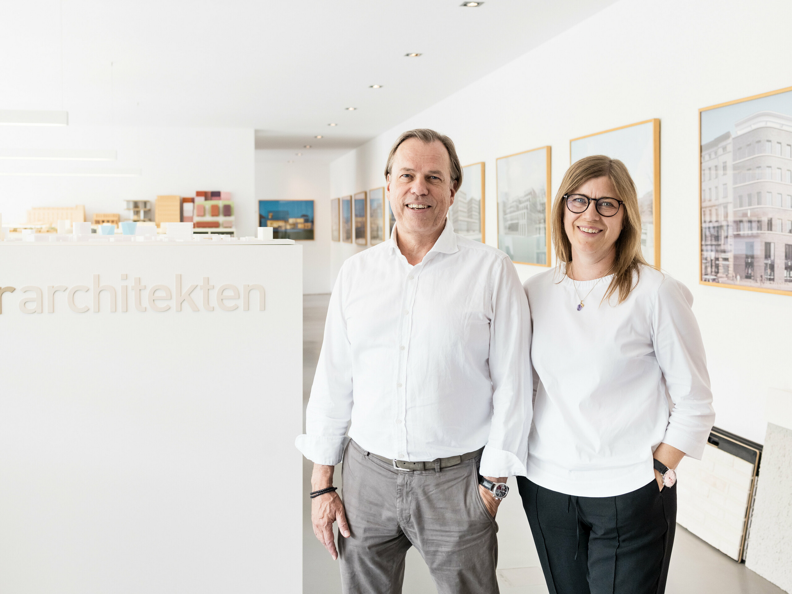 Leona und Andreas Geitner, die leitenden Architekten des Projektes in Ratingen (DE), stehen lächelnd vor einer weißen Wand mit dem Schriftzug "Geitner Architekten". Beide tragen weiße Hemden, was dem Bild ein professionelles und einheitliches Erscheinungsbild verleiht. Im Hintergrund sind Architekturmodelle und gerahmte Bilder von Gebäudeprojekten zu sehen, die das kreative und innovative Umfeld des Architekturbüros widerspiegeln. Die freundliche und einladende Atmosphäre unterstreicht die Professionalität und Kompetenz des Teams von Leona und Andreas Geitner.