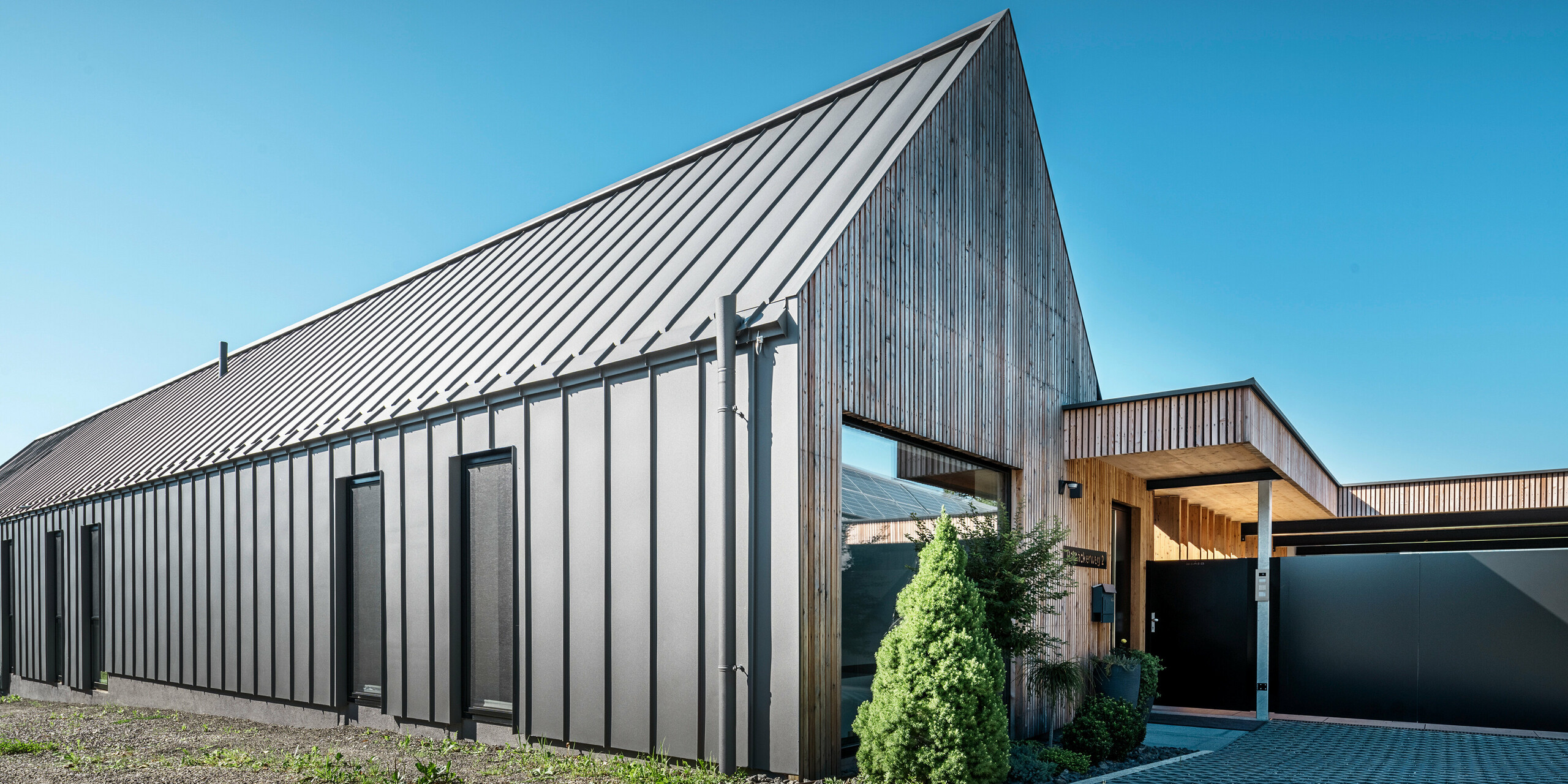 Eingangsbereich eines Einfamilienhauses mit angrenzender Garage in Pogier, Österreich, inmitten einer malerischen Landschaft. Dach und Fassade beider Gebäude sind mit langlebigem PREFALZ in dunkelgrau P.10 eingedeckt, was ein modernes Erscheinungsbild ergibt. Die Garage hat eine harmonisch integrierte Holzverkleidung, die sich nahtlos in die umgebende Landschaft einfügt. Der gepflasterte Hof und die klaren Linien des Entwurfs runden die Ästhetik des Grundstücks ab, während die sanften Hügel im Hintergrund das Bild einer ruhigen grünen Oase im grünen Herzen Österreichs vervollständigen.