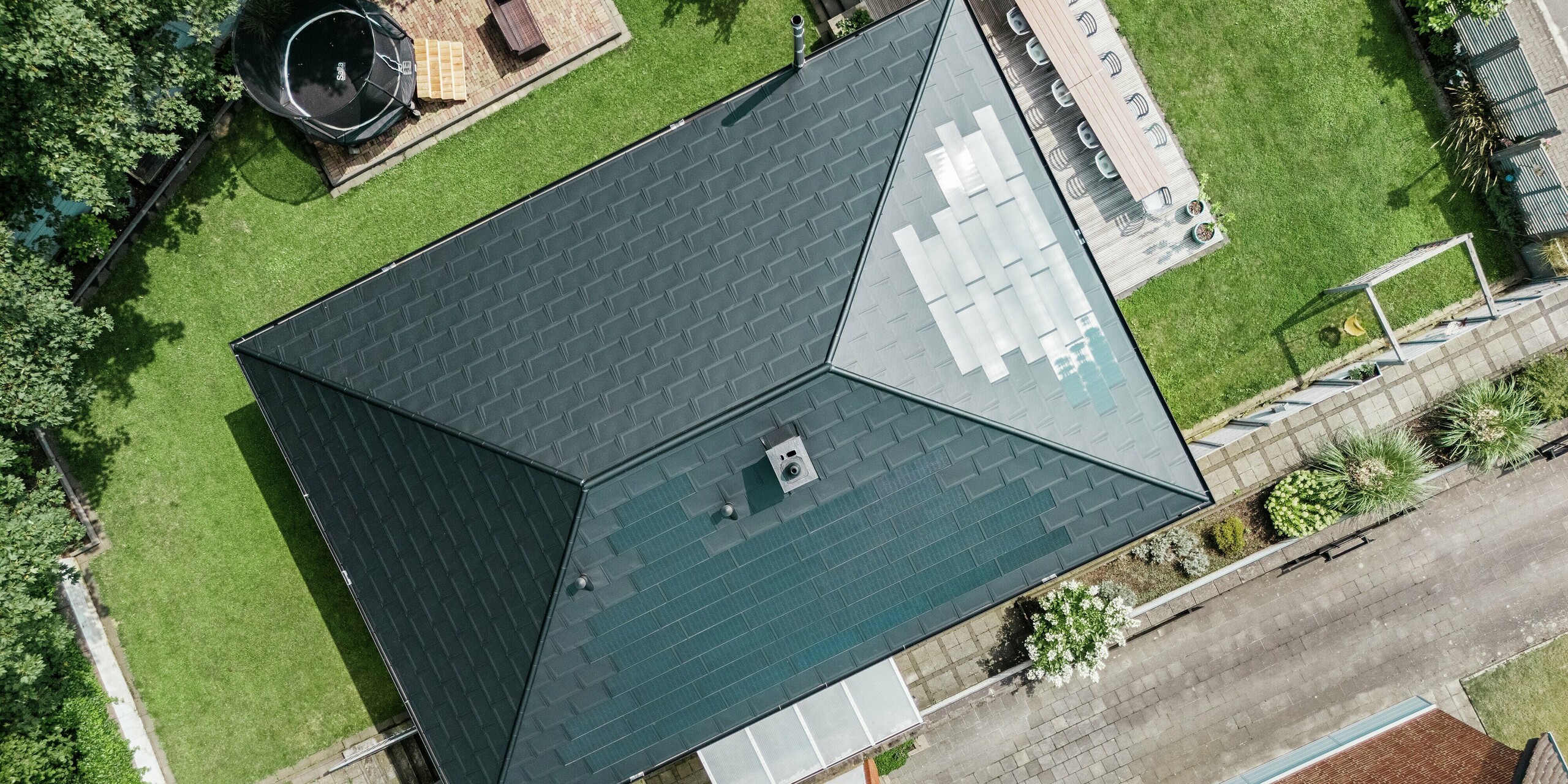 Vogelperspektive auf ein modernes PREFA Aluminiumdach eines Bungalows in Rommerskirchen. Die innovative Kombination aus Solardachplatten und Dachplatten R.16 in P.10 Schwarz sorgt für nachhaltige Energiegewinnung und optimalen Schutz. Das elegante Design fügt sich nahtlos in die grüne Umgebung ein und sorgt für einen harmonischen Gesamteindruck.