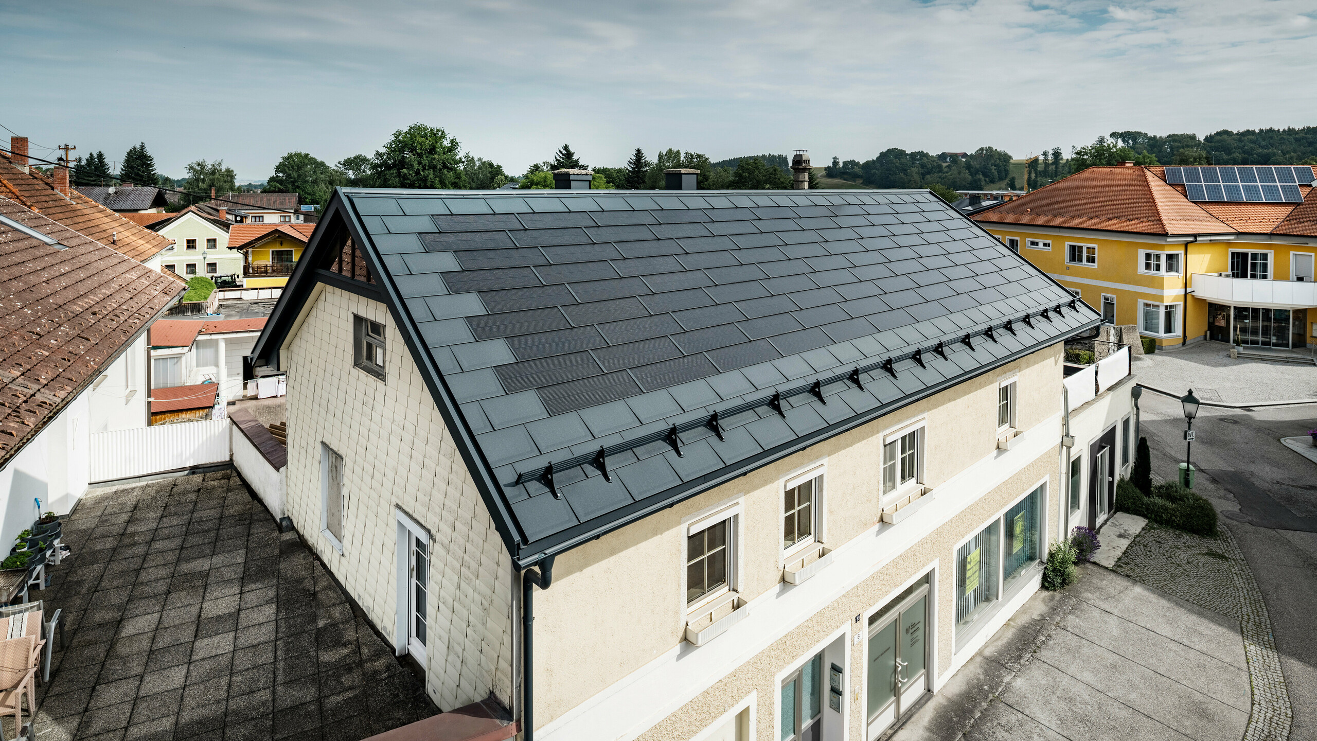 Vy från ovan över ett fristående hus i Mettmach, Österrike. Den äldre byggnaden har renoverats med antracitfärgade PREFA solar takplatta. Snöskyddssystemen och de klara linjerna på solcellstakpanelerna understryker det rena utförandet av PREFAs aluminiumprodukter. Det orangefärgade tegeltaket på grannbyggnaden kontrasterar i bakgrunden. Solcellsmodulerna är installerade på taket till grannhuset i en klassisk on roof-lösning, vilket visar på mångfalden av energieffektiv takdesign.