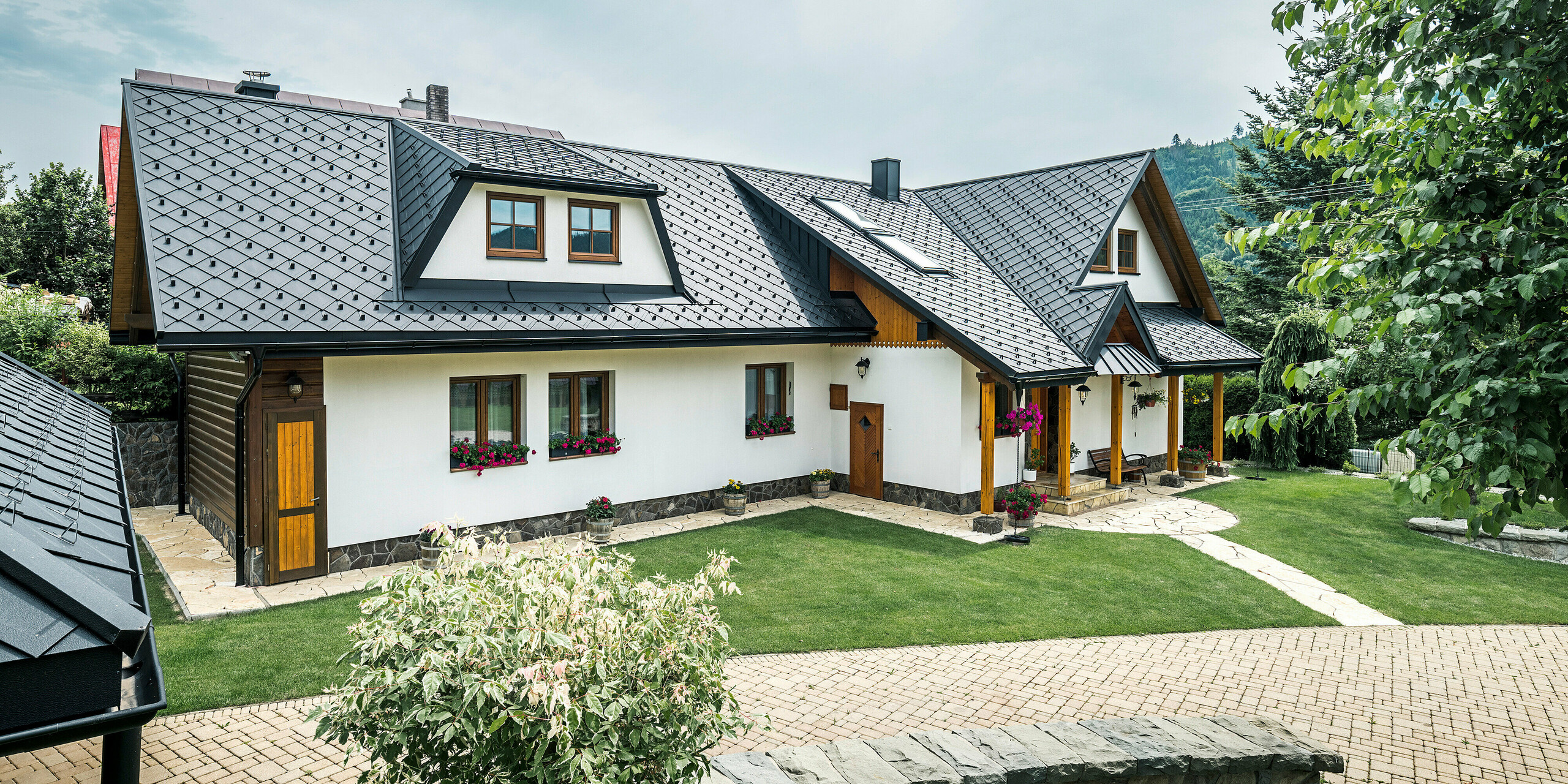 Ansicht eines traditionellen Einfamilienhauses in Stara Bystrica, Slowakei, mit einem Dach aus PREFA Dachrauten 29 × 29 in P.10 Schwarz. Die Aluminiumrauten ziehen sich gleichermaßen über die Trapezgaube auf der Vorderseite des Gebäudes. Im Vordergrund des Bildes ist eine gepflegte Gartenanlage zu sehen. Der Eingangsbereich ist mit einem Vordach geschützt, das mit PREFALZ verkleidet ist. Die sorgfältig gestaltete Einfahrt und die Blumenarrangements unterstreichen das einladende und stilvolle Gesamtbild.