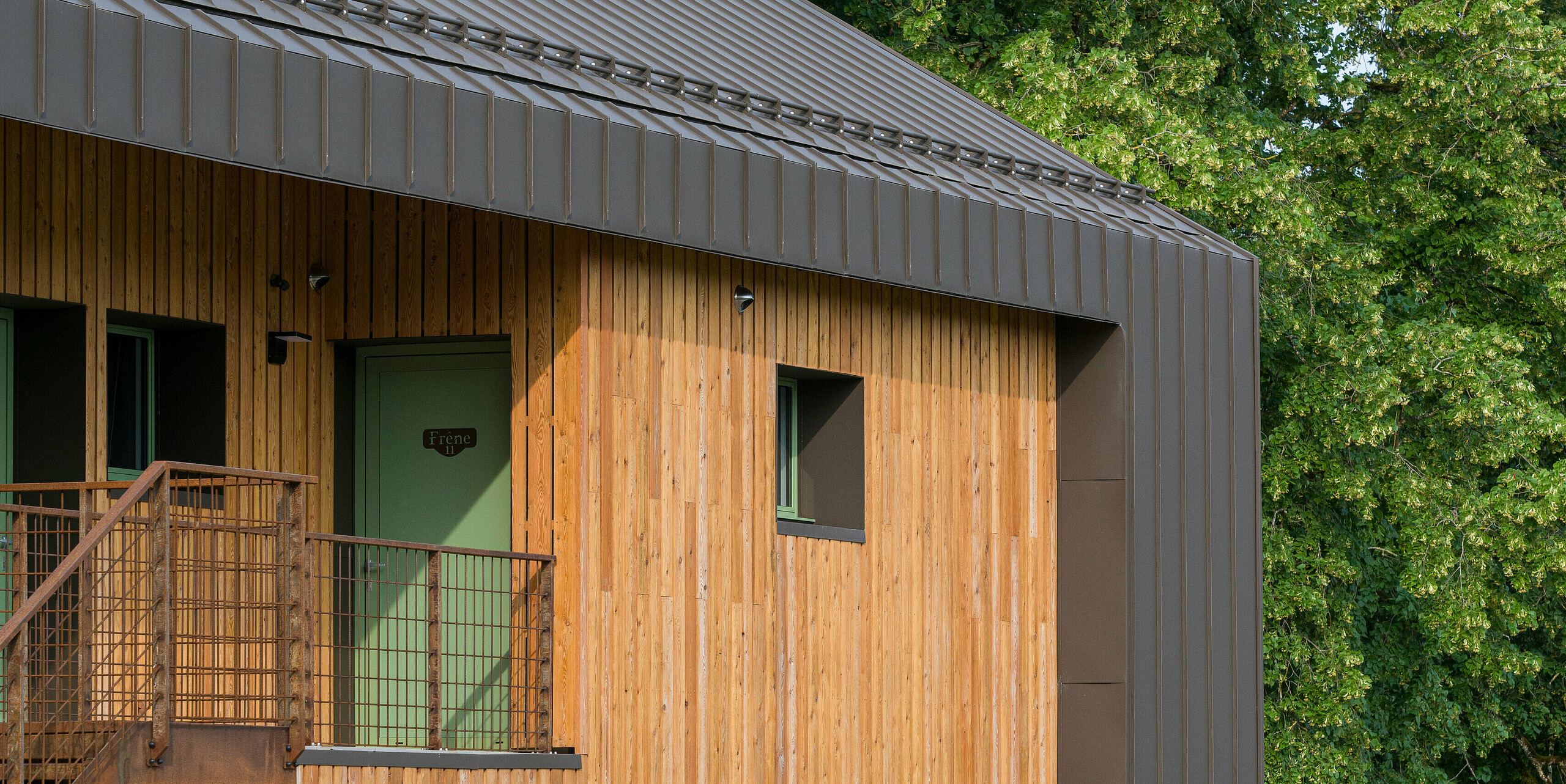 Seitliche Ansicht im Hochformat des Hôtel en Tilleul in Pont du Navoy (Frankreich). Das Gebäude verfügt über eine markante Holzfassade gepaart mit Aluminiumelementen bestehend aus PREFALZ in der Farbe P.10 Braun. Das Dach wurde vollständig mit einer Stehfalzeindeckung verkleidet, die sich über die Traufe zieht und auch die Ränder der Fassade schmückt. Vor dem Gebäude stehen vier Stühle und ein Holztisch. Das Stockwerk darüber ist mit einem Metallgeländer begrenzt. Der Hintergrund besteht aus großen Bäumen.