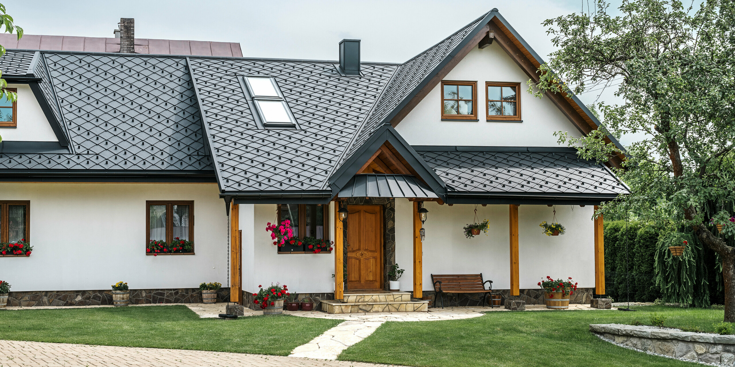 Eingangsbereich eines traditionellen Einfamilienhauses in Stara Bystrica, Slowakei, mit einem Aluminiumdach aus PREFA Dachrauten 29 × 29 in P.10 Schwarz. Das hochwertige Blechdach in eleganter Schuppenoptik umfasst Dachschrägenfenster und eine Veranda mit Vordach, das mit PREFALZ verkleidet ist. Die eleganten und zugleich robusten Aluminiumprodukte verleihen dem Haus ein zeitgemäßes und einladendes Erscheinungsbild. Der gepflegte Garten und die Blumenarrangements unterstreichen die gute Pflege des Gebäudes.