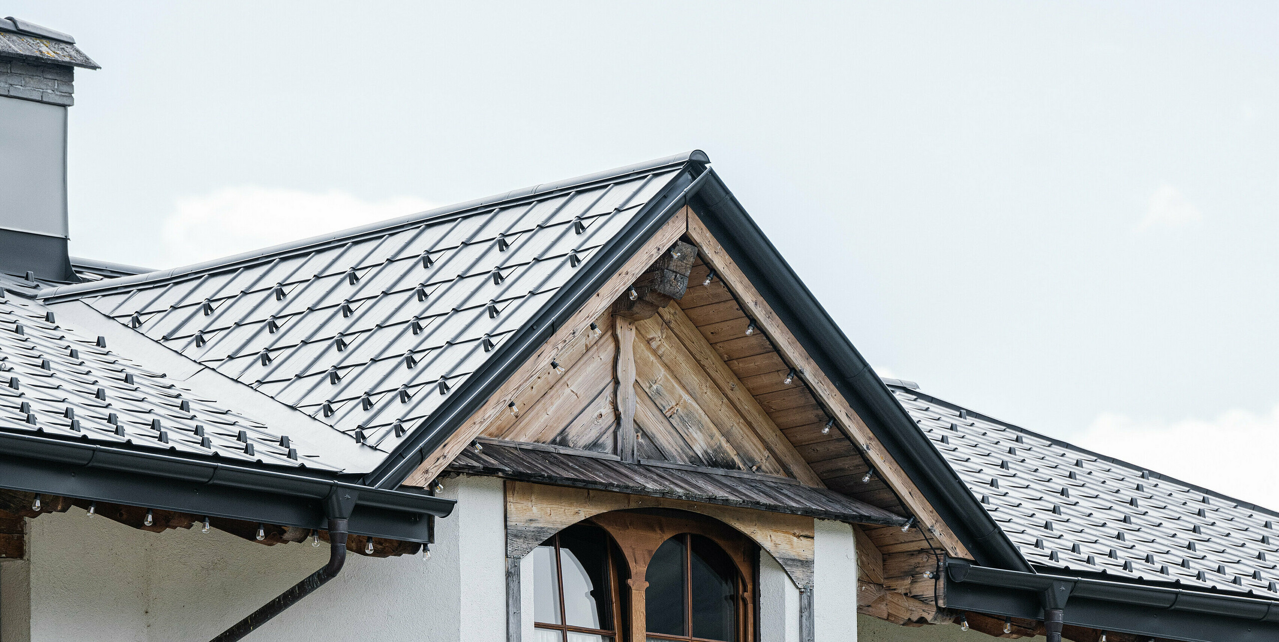 Eine Nahaufnahme eines traditionellen Alpenhauses in Auronzo di Cadore mit einem hellgrauen PREFA Dach, das die robuste Bauweise und den alpinen Stil der Region widerspiegelt. Sorgfältig verlegte Aluminiumfliesen passen zu den hölzernen Fensterläden und der verzierten Fassade, während langlebige Materialien das Haus vor den Elementen schützen und sich in das malerische Bergpanorama einfügen.