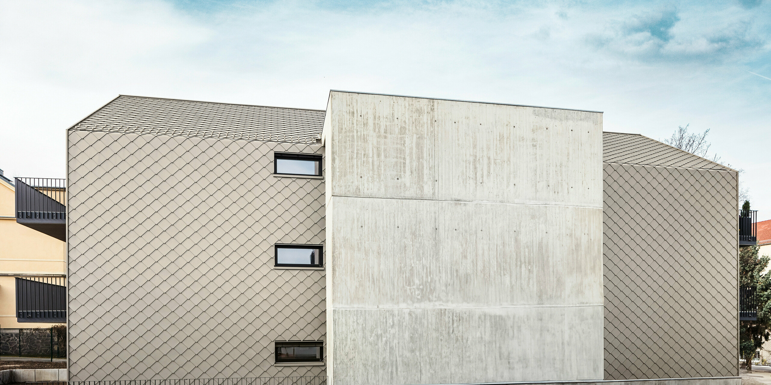 Seitenansicht eines Mehrfamilienhauses mit sechs Wohneinheiten in Dresden, Deutschland. Das Gebäude wird geschützt von einer Kombination aus PREFA Aluminium-Rauten 29 × 29 in Bronze und Sichtbeton. Das Bild zeigt die kontrastreiche Fassade, die aus einem Abschnitt in Beton besteht und einem Teil mit bronzefarbenen Aluminium-Rauten verkleidet wurde. Kleine Fenster und die klare Linienführung unterstreichen das minimalistische und moderne Design des Gebäudes.