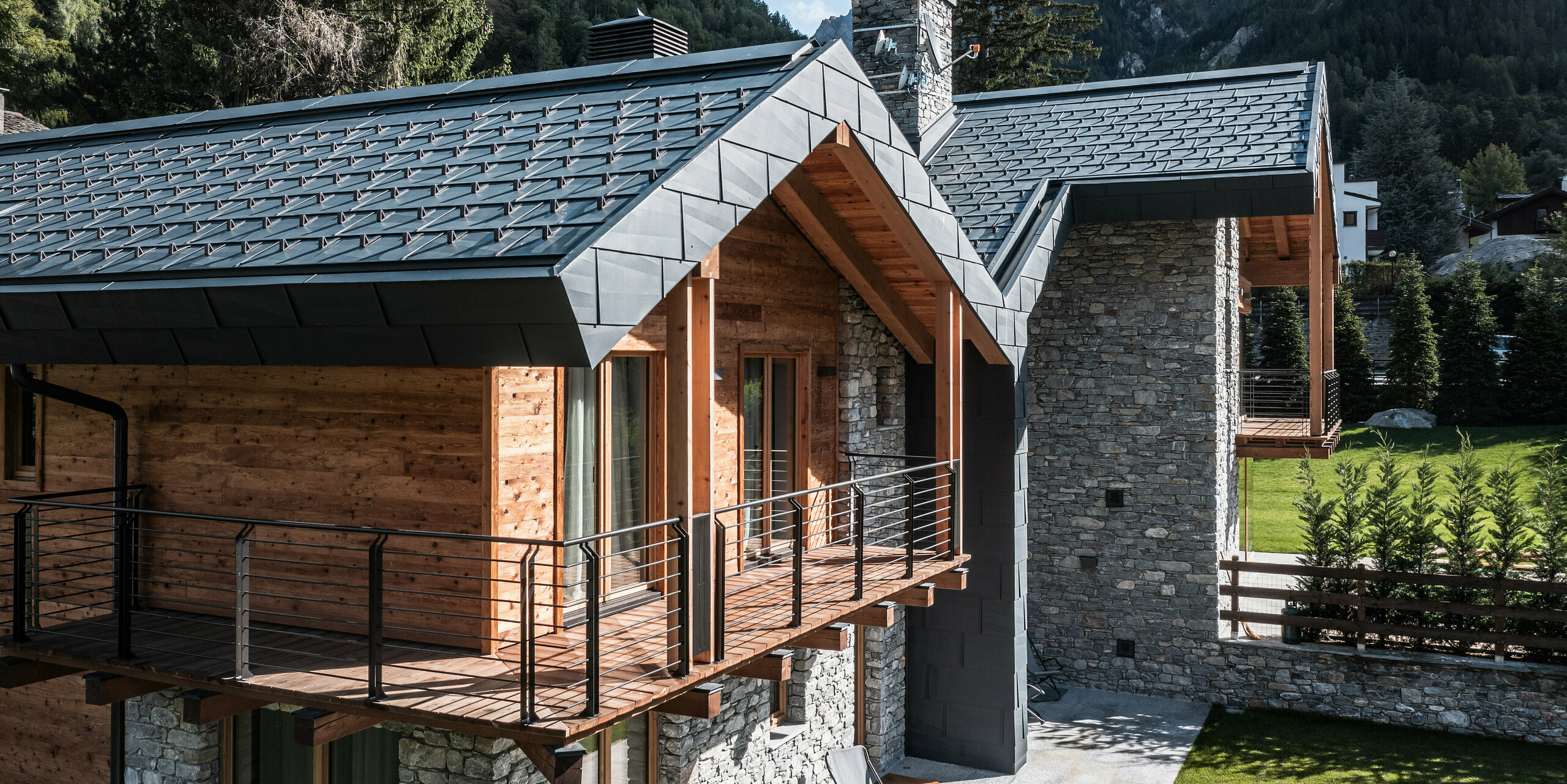 Moderne Mehrgenerationenvilla in Courmayeur mit einer beeindruckenden Gebäudehülle aus Stein, Holz und PREFA Aluminium. Die Dach- und Fassadenpaneele FX.12 in P.10 Steingrau fügen sich harmonisch in die alpine Landschaft des Aostatals ein. Die speziellen Paneele spielen dank ihrer unregelmäßigen Längs- und Querkantungen mit Licht und Schatten und erzeugen je nach Lichteinfall einzigartige optische Effekte. Im Vordergrund befindet sich ein großzügiger Balkon mit Edelstahlgeländer. Die beeindruckende Bergkulisse im Hintergrund unterstreicht das Spiel von Licht und Schatten auf den unregelmäßigen Dachkanten zusätzlich.