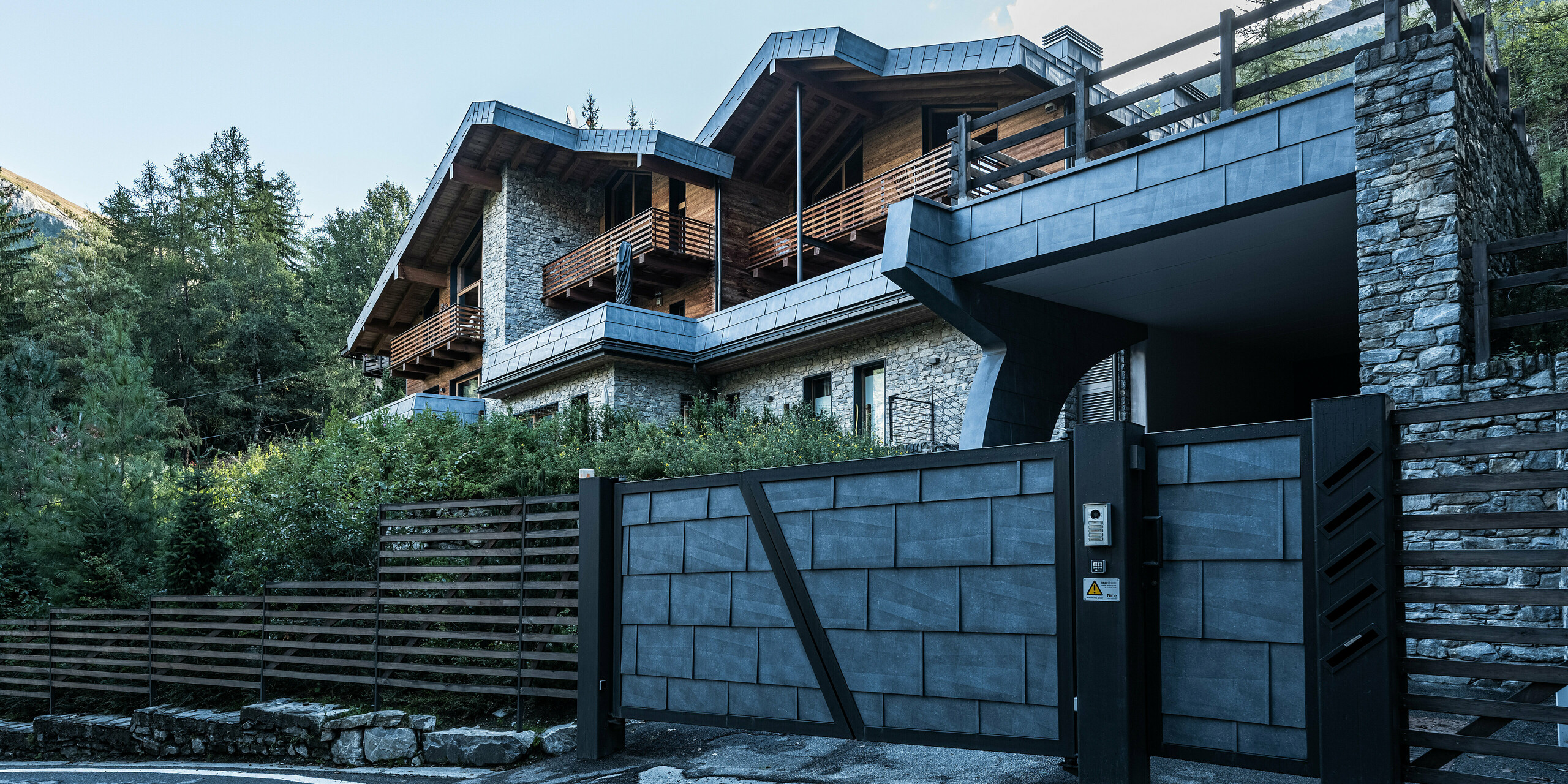 Eingangsbereich einer modernen Villa in Courmayeur, geprägt durch einen geschmackvollen Mix aus Stein-, Holz- und Aluminiumelementen. Die dynamische Dachkonstruktion aus schieferfarbenen PREFA FX.12 Dachplatten ergänzt die robusten Steinwände und warmen Holzbalkone und spiegelt den charakteristischen Alpenstil wider. Auch das hochwertige Eingangstor wurde mit robusten PREFA-Blechprodukten verkleidet. Die gepflegte Gartenanlage unterstreicht das luxuriöse Ambiente der exklusiven Residenz.