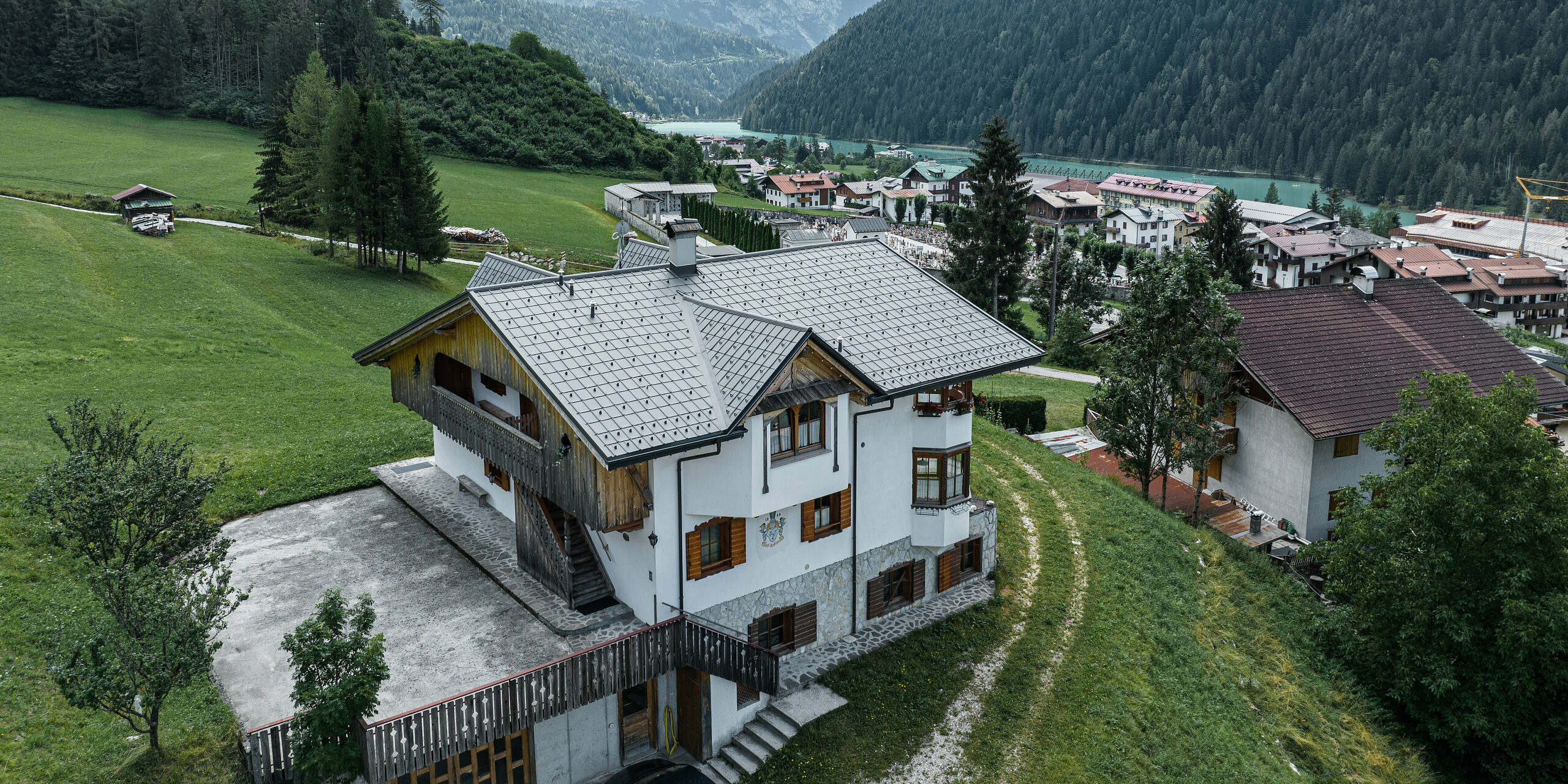 Luftaufnahme eines charmanten Bergchalets in Auronzo di Cadore, Italien, nach der Dachsanierung mit PREFA Aluminiumziegeln in Hellgrau. Das Chalet erhebt sich auf einer grünen Wiese mit einer beeindruckenden Bergkette im Hintergrund und zeigt ein malerisches Zusammenspiel von modernisierten Bauelementen und traditioneller Alpenarchitektur. Über dem Eingangsbereich ist eine Spitzgaube mit einem Holzgiebel zu sehen.