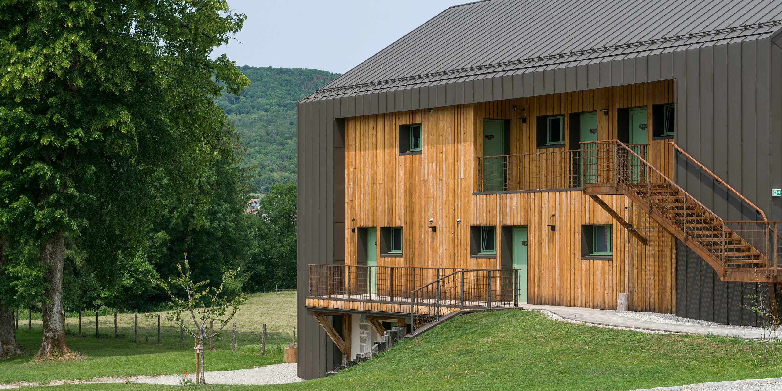 Seitliche Perspektive auf das Hôtel en Tilleul in Pont du Navoy (Frankreich). Das moderne Gebäude verfügt über eine stimmige Außenhülle aus Holz und Aluminium. Für die Aluminiumelemente am Dach und an den Rändern der Fassade kam PREFALZ in der Farbe P.10 Braun zum Einsatz. Das braune Aluminium fügt sich perfekt ein in der naturbelassenen Landschaft mit vielen Hügeln und Bäumen. Zudem ist das Material die ideale Ergänzung zu der hellen Holzfassade. Zahlreiche Fenster sowie ein Stiegenaufgang mit einem Metallgeländer zu den oberen Stockwerken zieren die sichtbare Seite des Gebäudes. 