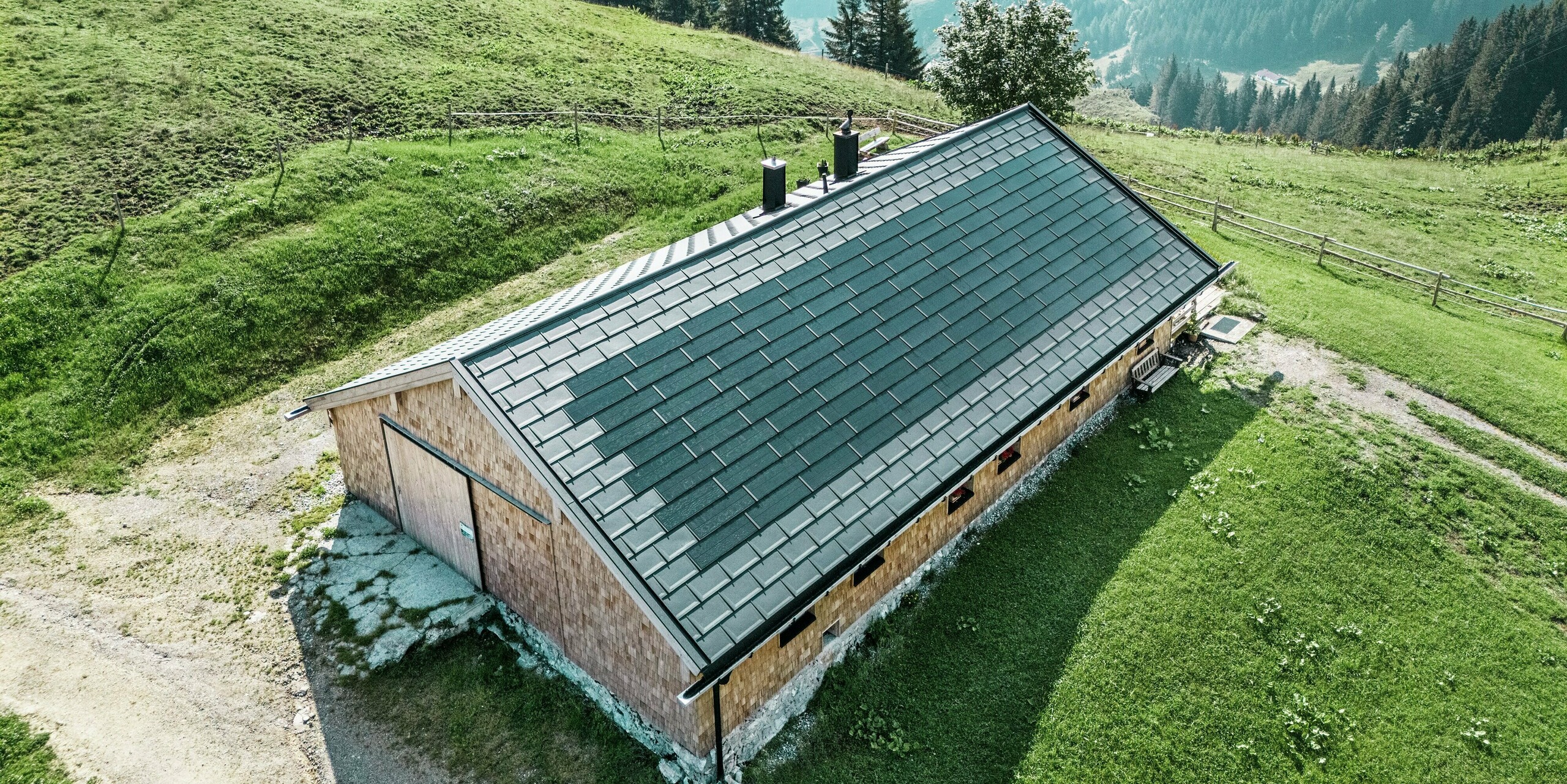 Drohnenaufnahme der Birkehütte auf der Walleralm in Bayrischzell inmitten einer idyllischen Berglandschaft. Das Dach wurde im Zuge einer Sanierung mit langlebigen PREFA Dachplatten R.16 in P.10 Schwarz und innovativen PREFA Solardachplatten ausgestattet. Diese moderne Lösung verbindet nachhaltige Energiegewinnung mit dem robusten Schutz bewährter PREFA Aluminiumprodukte und fügt sich harmonisch in die alpine Umgebung ein.