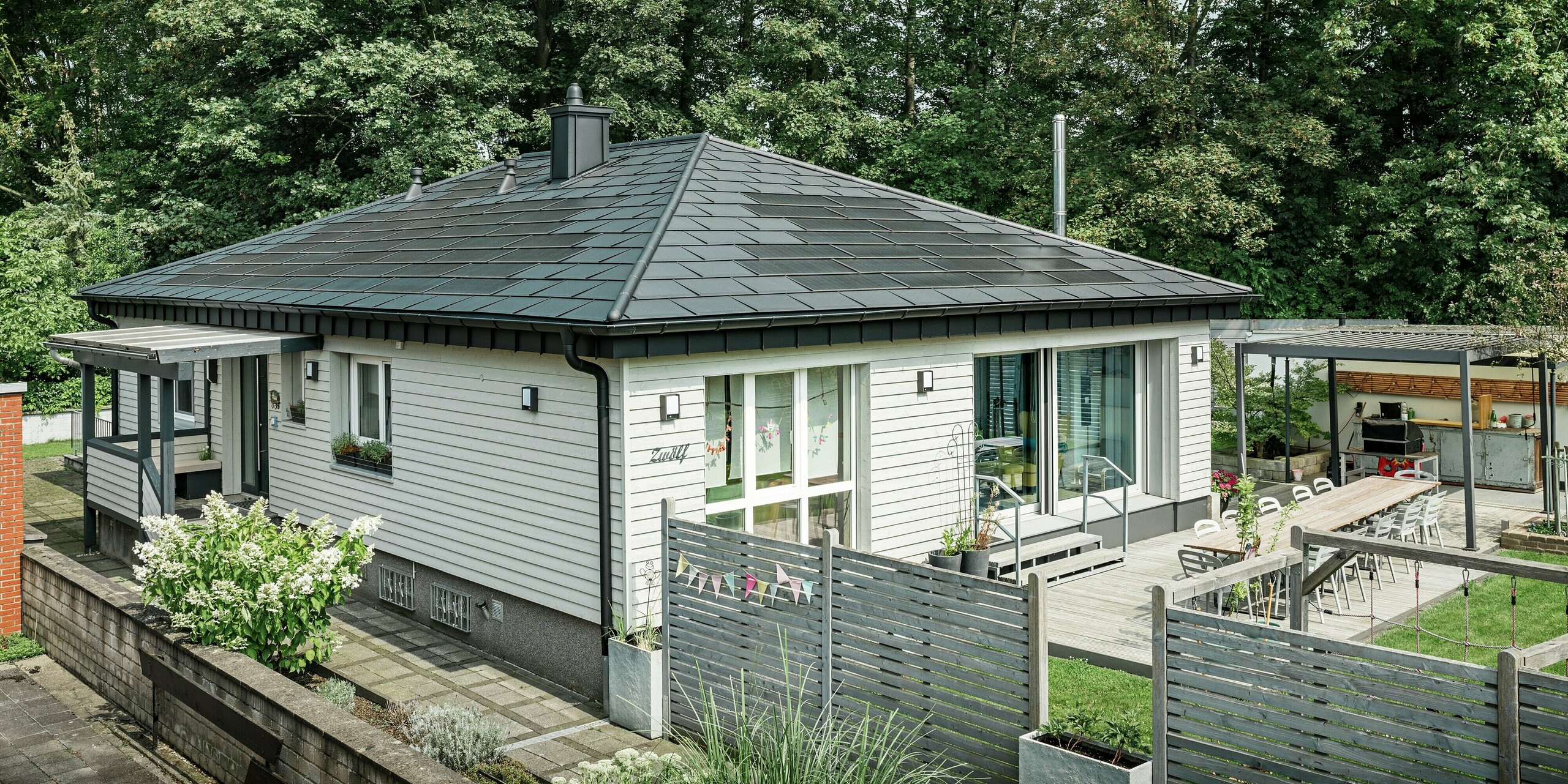 Ein Bungalow in Rommerskirchen (Deutschland) nach der Sanierung mit einem eleganten PREFA Aluminiumdach. Die innovative Kombination aus PREFA Solardachplatten und Dachplatten R.16 in P.10 Schwarz bietet dauerhaften Schutz bei gleichzeitiger Energiegewinnung. Perfekt integriert sind Saumrinne und Ablaufrohr, die für eine effiziente Dachentwässerung in hochwertigem Design sorgen. Der Zugang zum Einfamilienhaus ist mit einem Holzzaun gesichert. Zwischen den Zaunelementen befinden sich niedrige Beete, die den Blick auf Garten und Terrasse freigeben. 