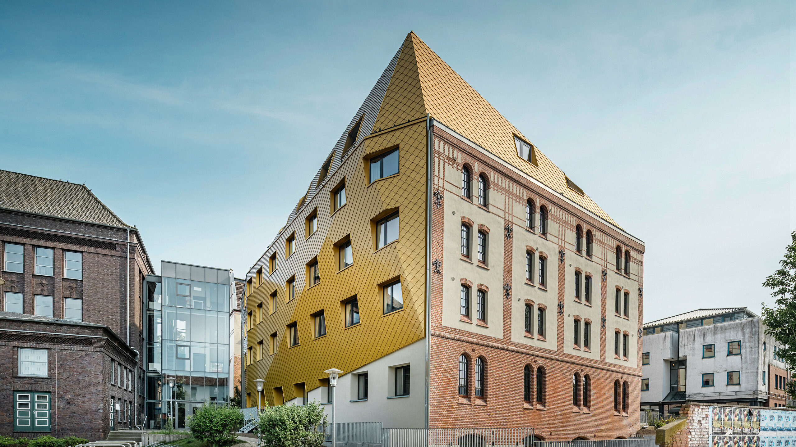Golvbild av "The Station" i Kiel. Studentbostaden syns diagonalt från sidan. Bilden visar tydligt den slående kontrasten mellan aluminiumfasaden av PREFA tak- och väggromboider 29 × 29 i Mayagold till vänster och den kulturminnesmärkta puts- och tegelfasaden till höger.