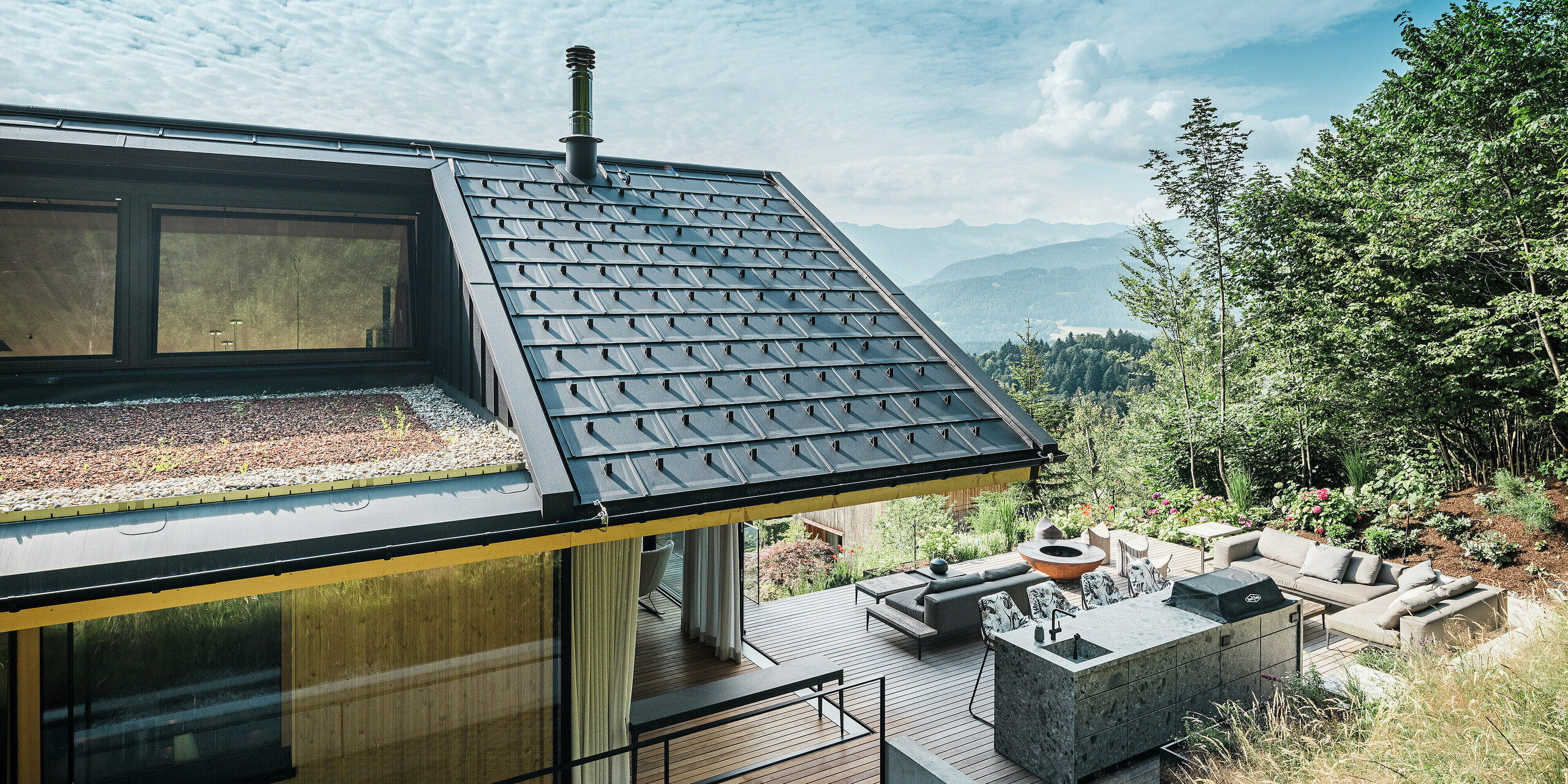 Blick auf die Terrasse eines Einfamilienhauses in Egg, Vorarlberg, mit PREFA Dachplatten R.16 in P.10 Schwarz, Schneefangsystem und Dachentwässerung. Das moderne Aluminiumdach verbindet Langlebigkeit mit ästhetischer Eleganz. Im Vordergrund die großzügige Terrasse mit Außenküche und stilvollen Gartenmöbeln, eingebettet in die natürliche Umgebung mit dem atemberaubenden Bergpanorama im Hintergrund.