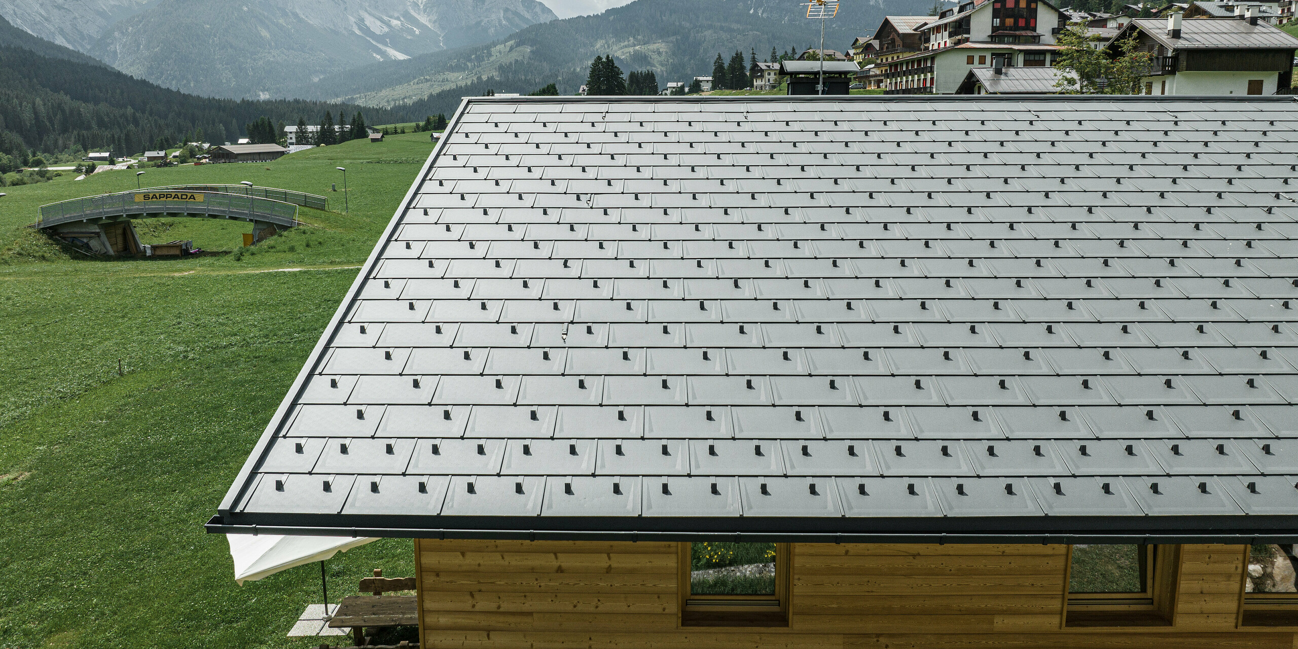 Dachansicht eines Einfamilienhauses in Sappada, Italien, umgeben von der majestätischen alpinen Kulisse. Das Haus ist mit PREFA Dachplatten R.16 in P.10 Schwarz ausgestattet, die ein modernes, lineares Muster bilden und einen ansprechenden Kontrast zur natürlichen Holzfassade darstellen. Diese hochwertigen, widerstandsfähigen Aluminiumdachplatten bieten einen langfristigen Schutz und unterstreichen das Engagement von PREFA für Ästhetik und Funktionalität.