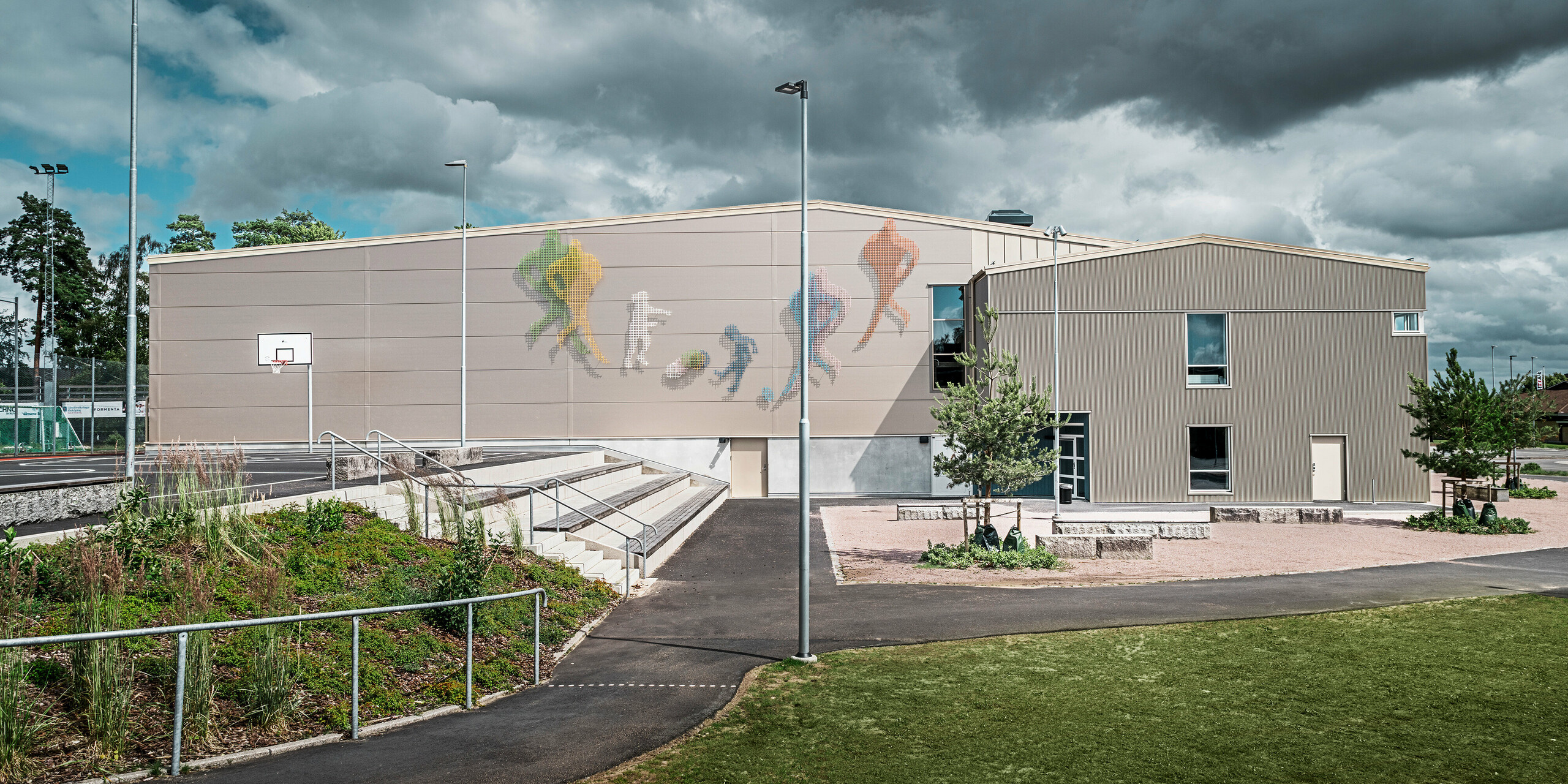 Außenansicht der Park Arena in Värnamo, Schweden, mit einem farbenfrohen Wandkunstwerk, das sportliche Figuren zeigt. Die Fassade ist mit dem PREFA Zackenprofil in Bronze verkleidet, das für eine moderne und langlebige Gebäudehülle sorgt. Im Vordergrund sind der Pausenbereich und ein Basketballplatz zu sehen, die den sportlichen Charakter der Anlage unterstreichen.