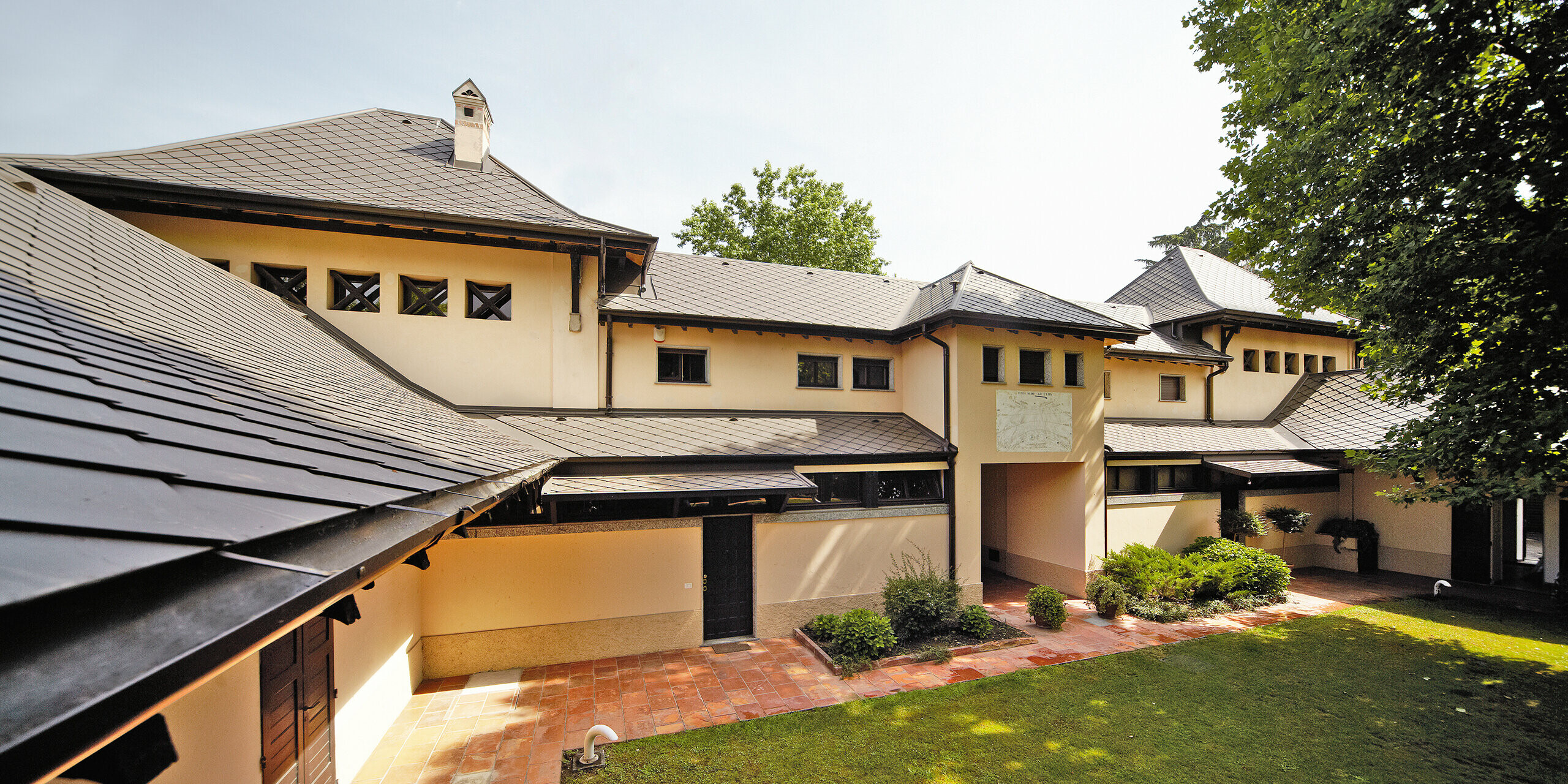 Italiensk villa med rombtak i P.10 brun från PREFA med beige fasadfärg och skuggig innergård