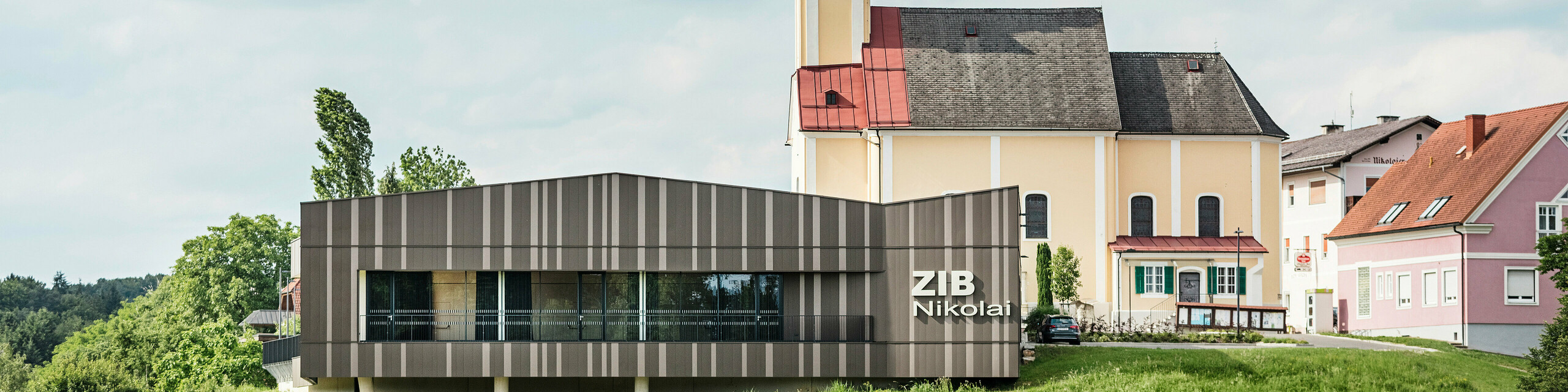 Das Veranstaltungszentrum ZIB Nikolai in St. Veit in der Südsteiermark mit einer modernen, dynamischen Aluminiumfassade in braun-beigegrauen Tönen, im Vordergrund eine grüne Wiese, im Hintergrund die Kirche von St. Nikolai mit gelber Fassade und rotem Dach.