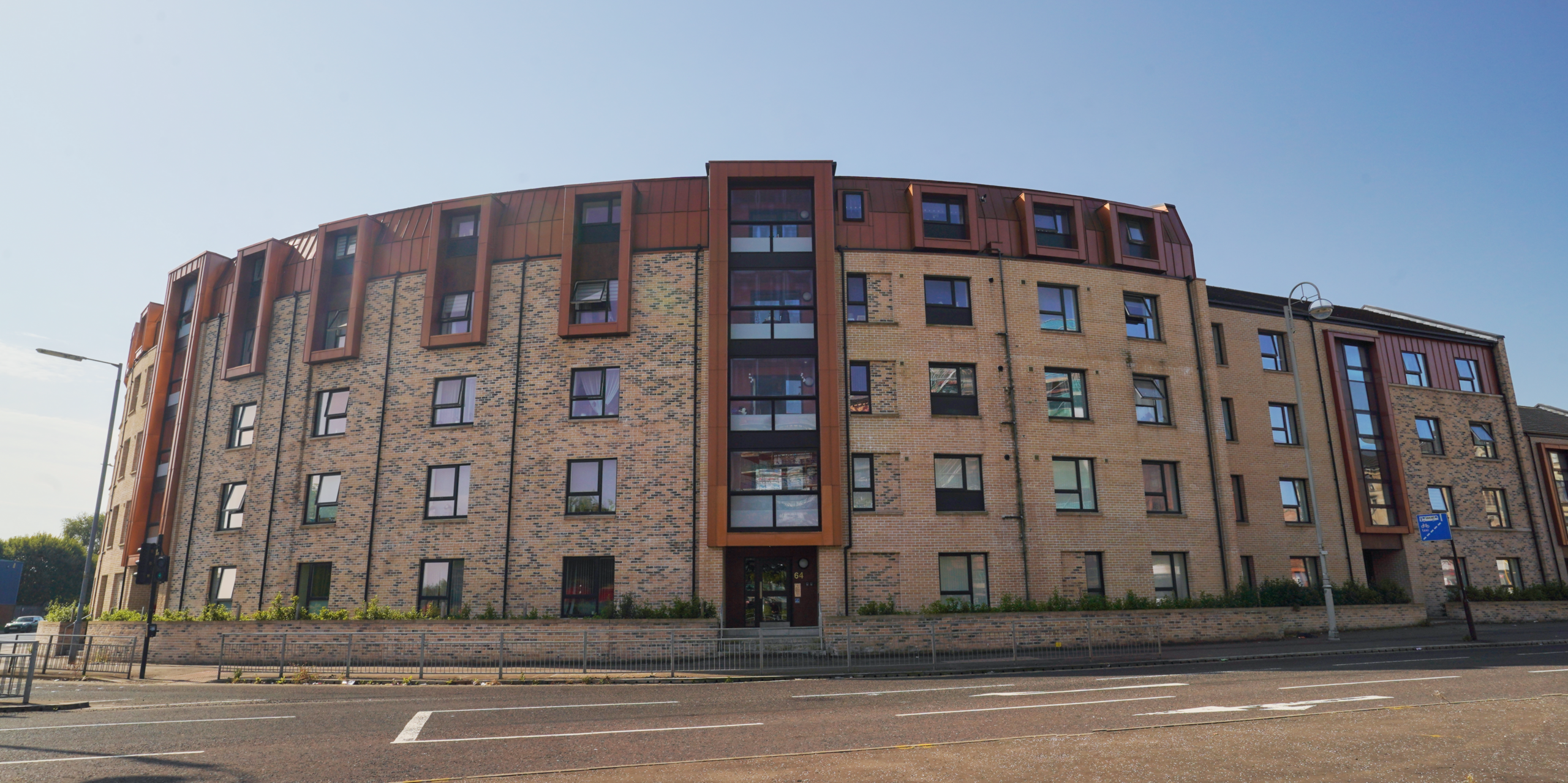 Blick von der Straße auf eine moderne Wohnhausanlage in der Nethan Street in Glasgow. Die Wohnalage sticht durch die Verwendung von rund 500m² FALZONAL in der Farbe Neukupfer an Dach und Fassadenteilen hervor. Die warme Kupferfarbe des Aluminiums ergänzt die Ziegelstruktur des Gebäudes und verleiht dem Objekt eine zeitgemäße und anspruchsvolle Ausstrahlung. Die harmonische Integration von Farbe und Material unterstreicht das Bestreben, städtischen Wohnraum ästhetisch und funktionell zu gestalten.