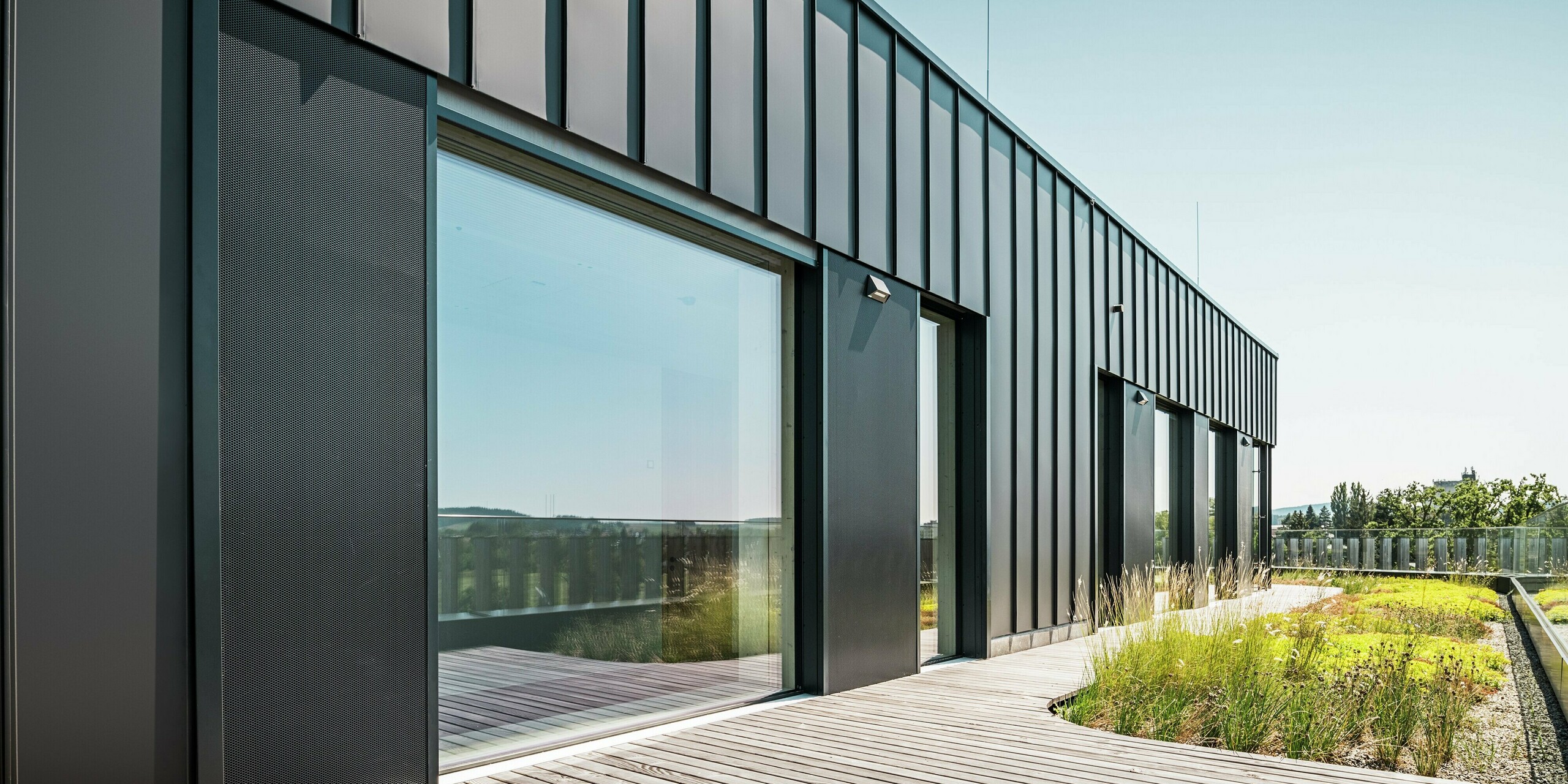 Die Dachterrasse der Team 7 Welt in Ried im Innkreis kombiniert eine PREFALZ Fassade in Schwarzbraun mit großen Glasfronten und einer begrünten Umgebung. Die hochwertige Aluminiumverkleidung bietet Langlebigkeit und modernen Stil, während die natürlichen Elemente des Gründachs für ein harmonisches und nachhaltiges Design sorgen. Perfekt für zeitgemäße Architektur mit ökologischer Ausrichtung.