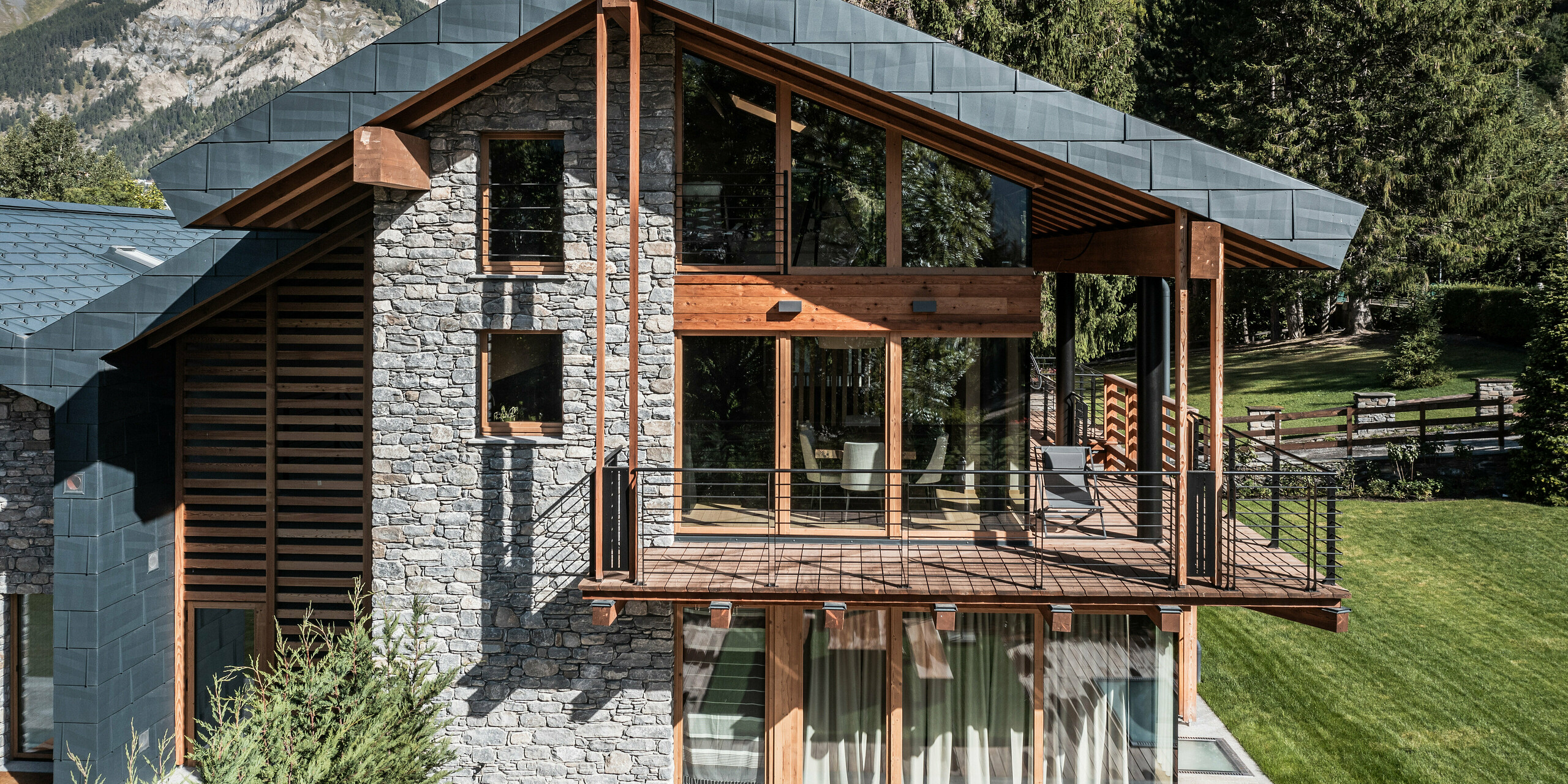 Luxusvilla in Courmayeur mit einer auffälligen Kombination aus Holz, Stein und PREFA Aluminium in Form von Dach- und Fassadenplatten FX.12 in P.10 Steingrau. Die strukturierte Fassadenverkleidung und die großen Fensterfronten bieten eine natürliche Ästhetik. Ein großer überdachter Holzbalkon mit Blick auf die alpine Landschaft bietet eine atemberaubende Aussicht. Das sorgfältige Design fügt sich nahtlos in die umliegende Bergkulisse ein und schafft eine dramatische Kulisse.