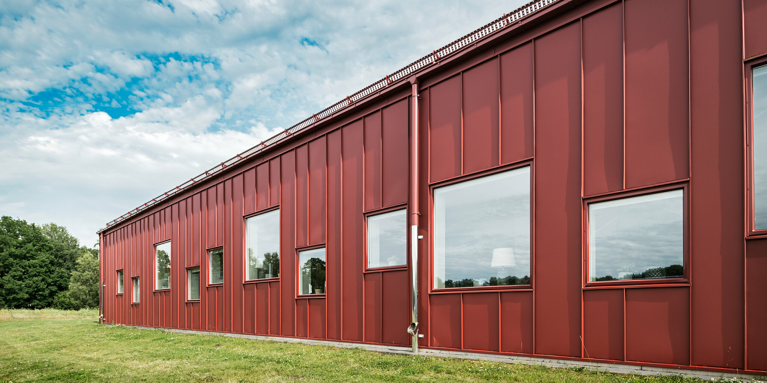 Die Rückseite der Schulkantine Gröna Hjärtat in Broby, Schweden, zeigt eine moderne und zugleich robuste Fassade aus PREFALZ in Oxydrot. Die langlebige Aluminiumverkleidung unterstreicht die hohe Qualität und Beständigkeit der Konstruktion. Das Dachentwässerungssystem ist ebenfalls in Oxydrot ausgeführt, was das einheitliche Erscheinungsbild verstärkt und die ästhetische Harmonie der Architektur betont.