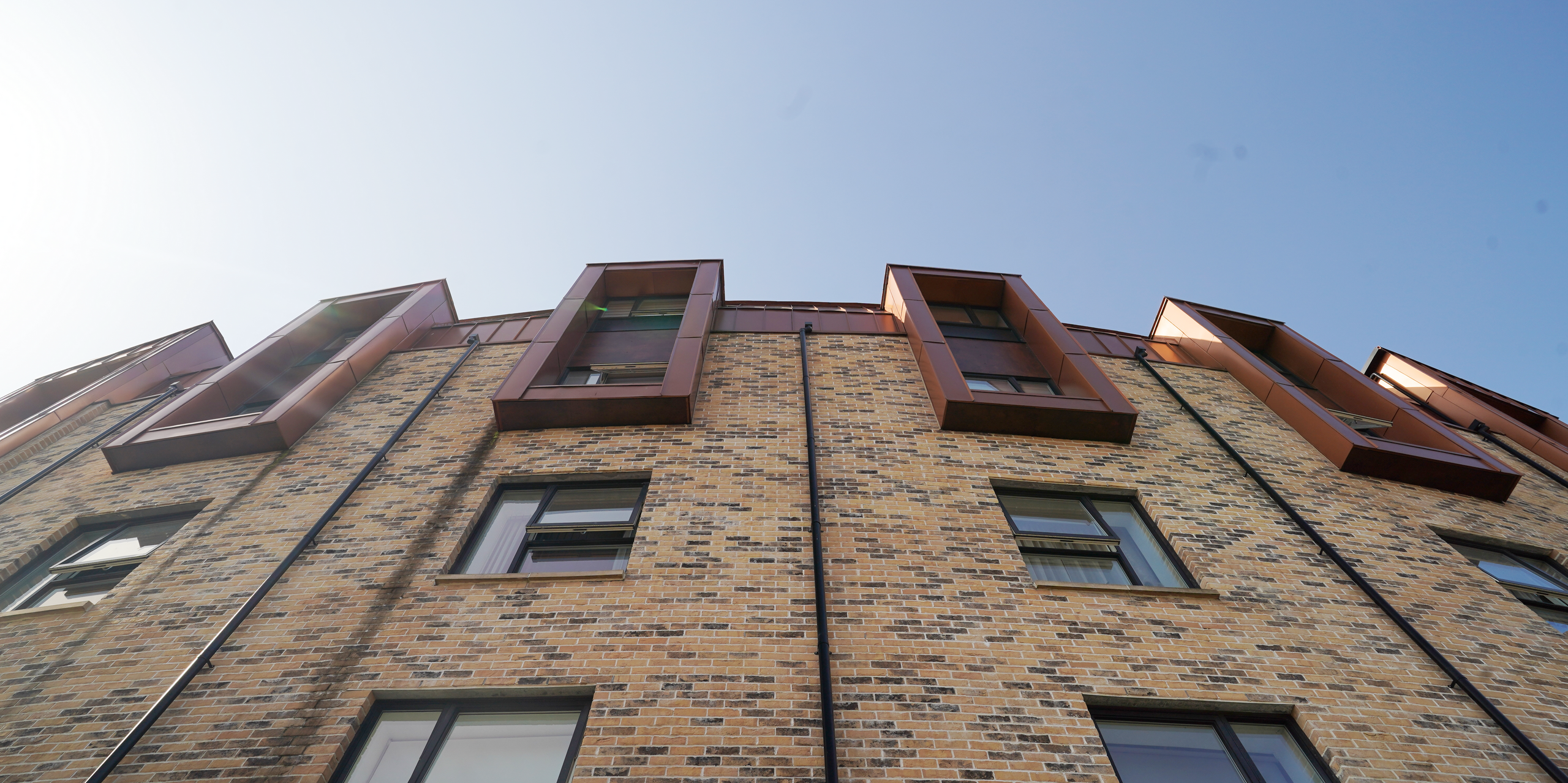 Aufwärtsblick auf die Fassade einer Wohnhausanlage in der Nethan Street in Glasgow, die sich durch die Verwendung von FALZONAL in Neukupfer auszeichnet, einem langlebigen und robusten PREFA Aluminiumprodukt. Die vertikale Ausrichtung der Fenster, die mit dem kupferfarbenen Aluminium umrahmt wurden, unterbricht die horizontale Gliederung der Ziegelmauer und verleiht dem Gebäude eine dynamische und moderne Ausstrahlung. Die Qualität und Farbgebung des FALZONAL fügt sich nahtlos in die städtische Architektur ein und bietet gleichzeitig eine beständige und ästhetische Lösung für moderne Wohnkomplexe. Zwischen den Fenstern sind schwarze Ablaufrohre zur Dachentwässerung platziert worden, die in dieser Perspektive die Rundung des Bauwerks verdeutlichen.