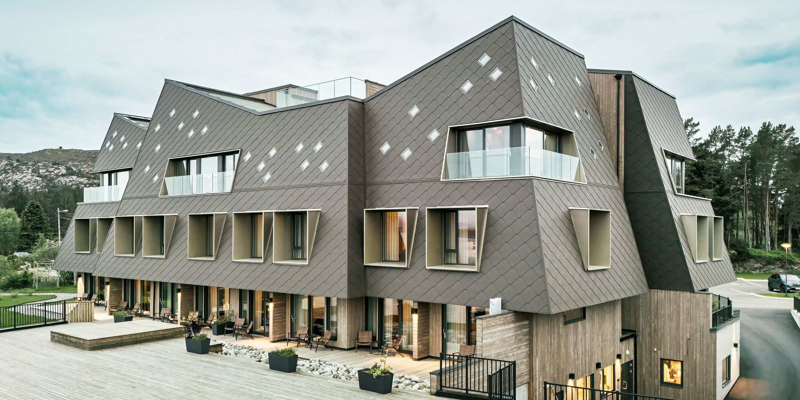 Blick auf die Terrasse des Hotels Beckerwyc House in Bekkjarvik, Norwegen. Das moderne Gebäude wird von einer einer markanten Aluminiumfassade aus PREFA Wandrauten 44 × 44 in Nussbraun geschmückt. Die futuristische Architektur mit schrägen Dachflächen und hervortretenden Fensterelementen wird durch die großzügigen Außenbereiche ergänzt. Die natürliche Farbgebung der Fassade passt sich harmonisch in die umgebende Landschaft ein und unterstreicht das zeitgemäße Design des Gebäudes.