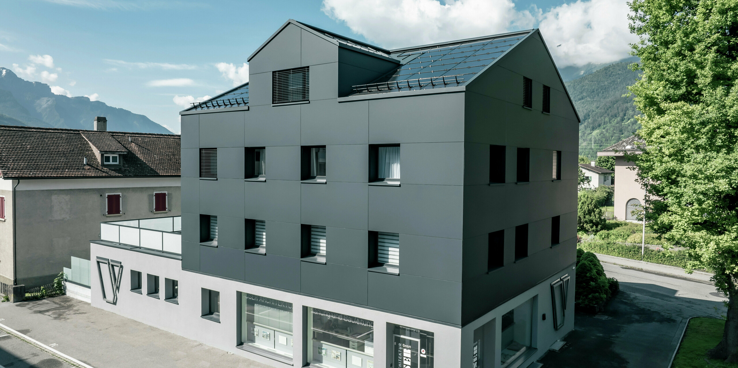 Modernisiertes Wohn- und Geschäftsgebäude nach der Fassadensanierung mit PREFABOND Aluminium Verbundplatten in Anthrazit. Die klare, elegante Linienführung der neuen Fassade verleiht dem Gebäude ein modernes Erscheinungsbild. Im Erdgeschoss befindet sich das Architekturbüro Voser. Die Umgebung mit Bäumen und Bergen rundet die harmonische Optik ab.