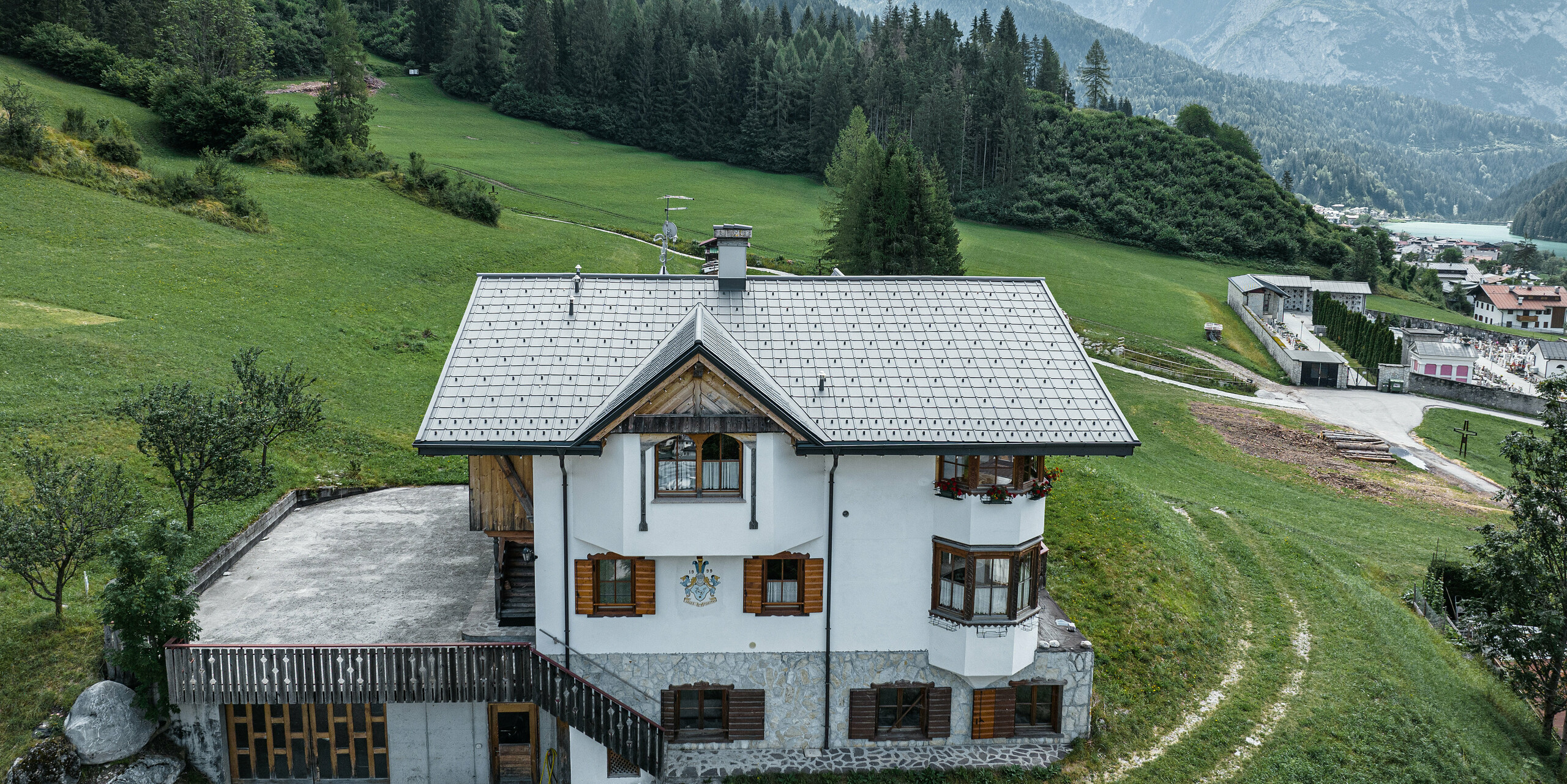 Blick aus der Vogelperspektive auf den Eingang der traditionellen Berghütte in Auronzo di Cadore nach der Dachsanierung mit PREFA Ziegeln aus Aluminium in P.10 Hellgrau. Das Gebäude mit seinem charakteristischen alpinen Charme und dem modernen Blechdach fügt sich harmonisch in die üppig grüne Landschaft und das beeindruckende Bergpanorama im Hintergrund ein und demonstriert eine gelungene Kombination aus Funktionalität und regionaler Architektur.